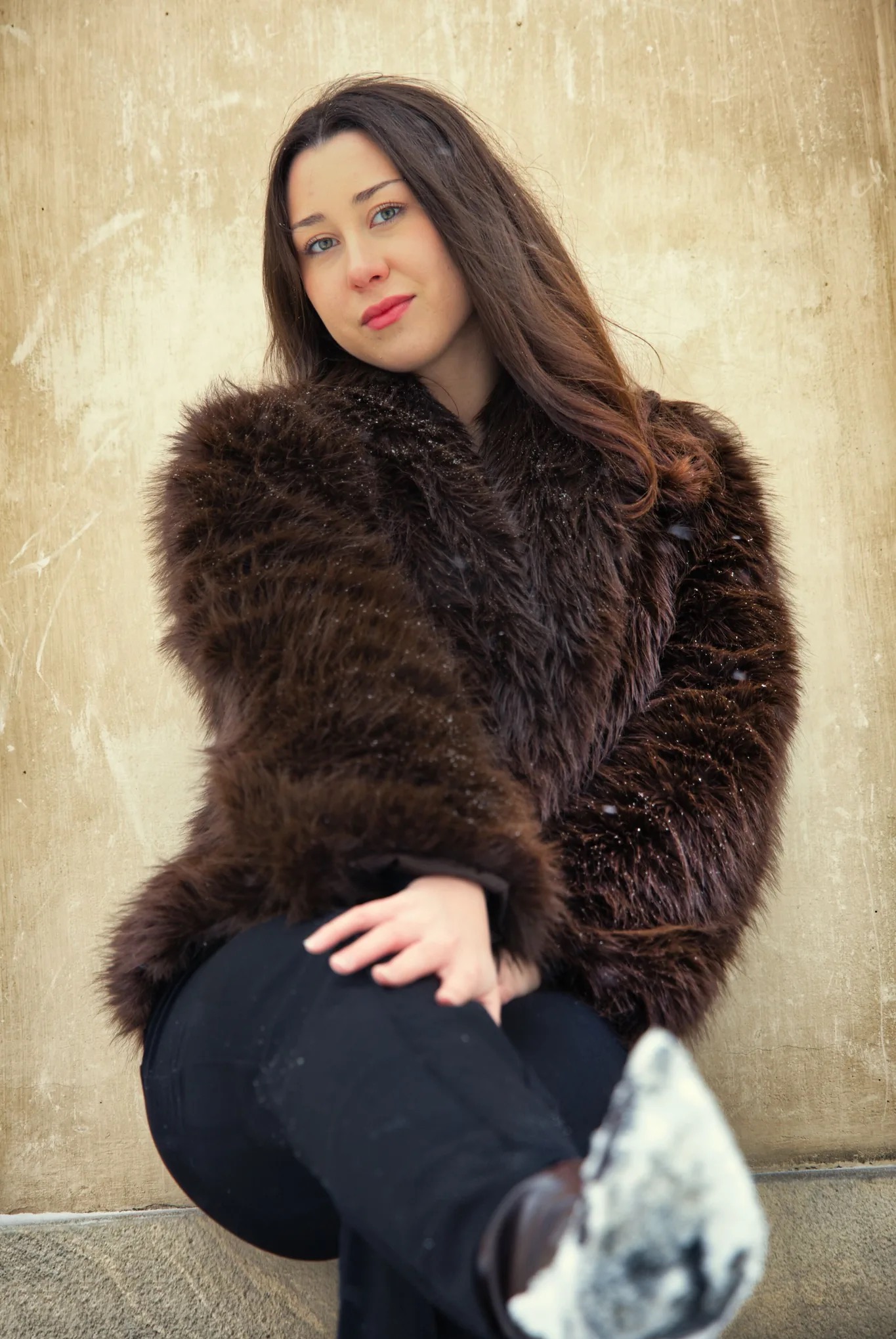 Portret kobiety w brązowym futrze na tle beżowej ściany. Uśmiechnięta brunetka z niebieskimi oczami pozuje, opierając się o ścianę. Widoczne płatki śniegu na futrze.