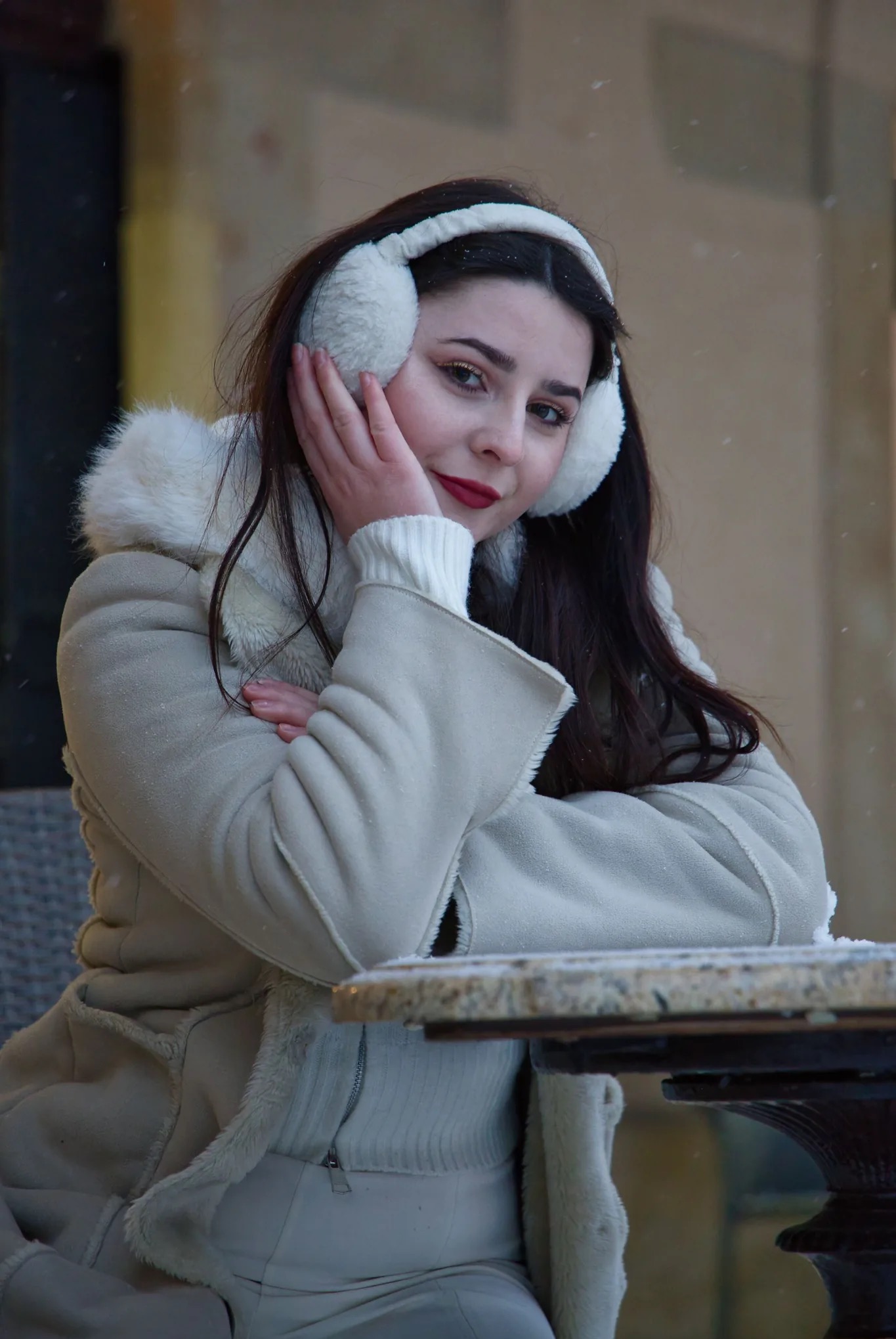 Portret kobiety w zimowej stylizacji: białe nauszniki, jasny płaszcz z futerkiem, czerwone usta. Delikatny uśmiech, śnieg pada w tle. Ujęcie z bliska, skupione na twarzy.