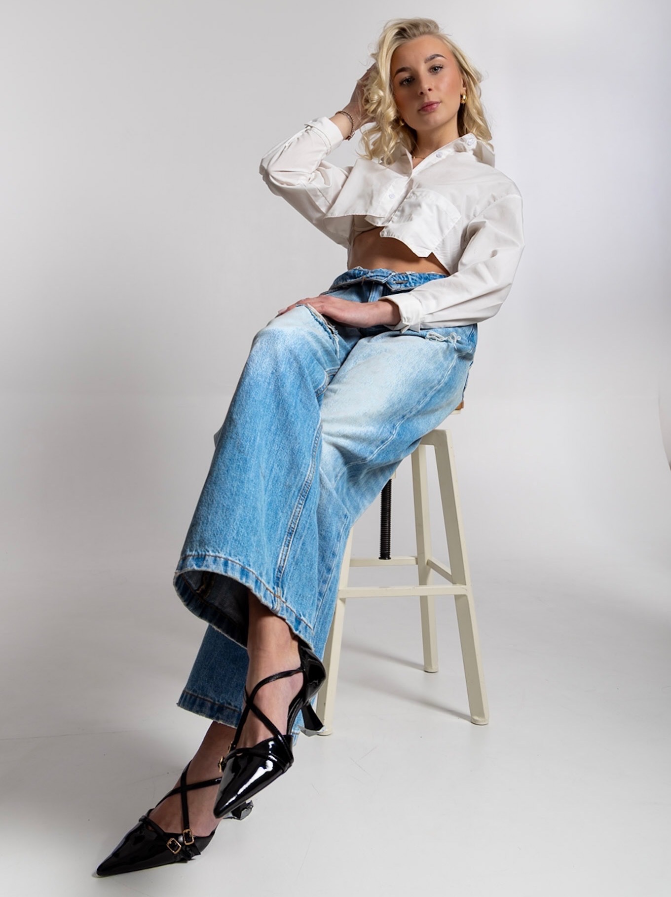 Blondwłosa modelka w białej koszuli i szerokich jeansach siedzi na krześle na jasnym tle, prezentując stylizację i pozę. Uwagę przyciągają czarne, lakierowane buty.