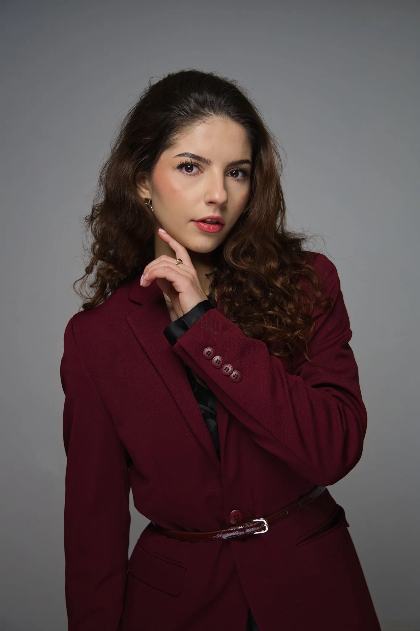 Portret kobiety w bordowym garniturze, z ręką przy twarzy, na szarym tle. Uwagę przyciąga wyrazisty makijaż i delikatna biżuteria. Ujęcie studyjne, profesjonalne.