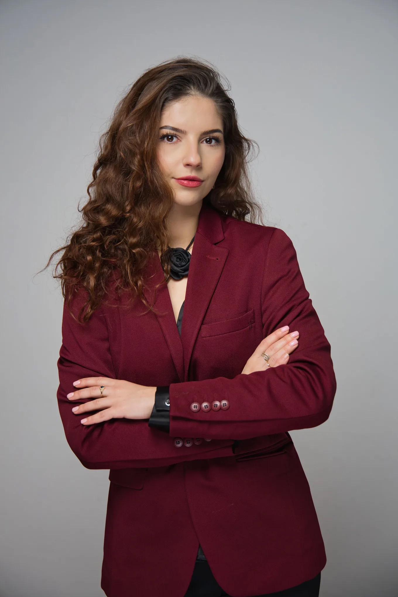 Portret kobiety w bordowej marynarce z rękami skrzyżowanymi na tle szarości. Sesja wizerunkowa w Krakowie. Widoczny pierścionek i ozdobny kwiat przy szyi.