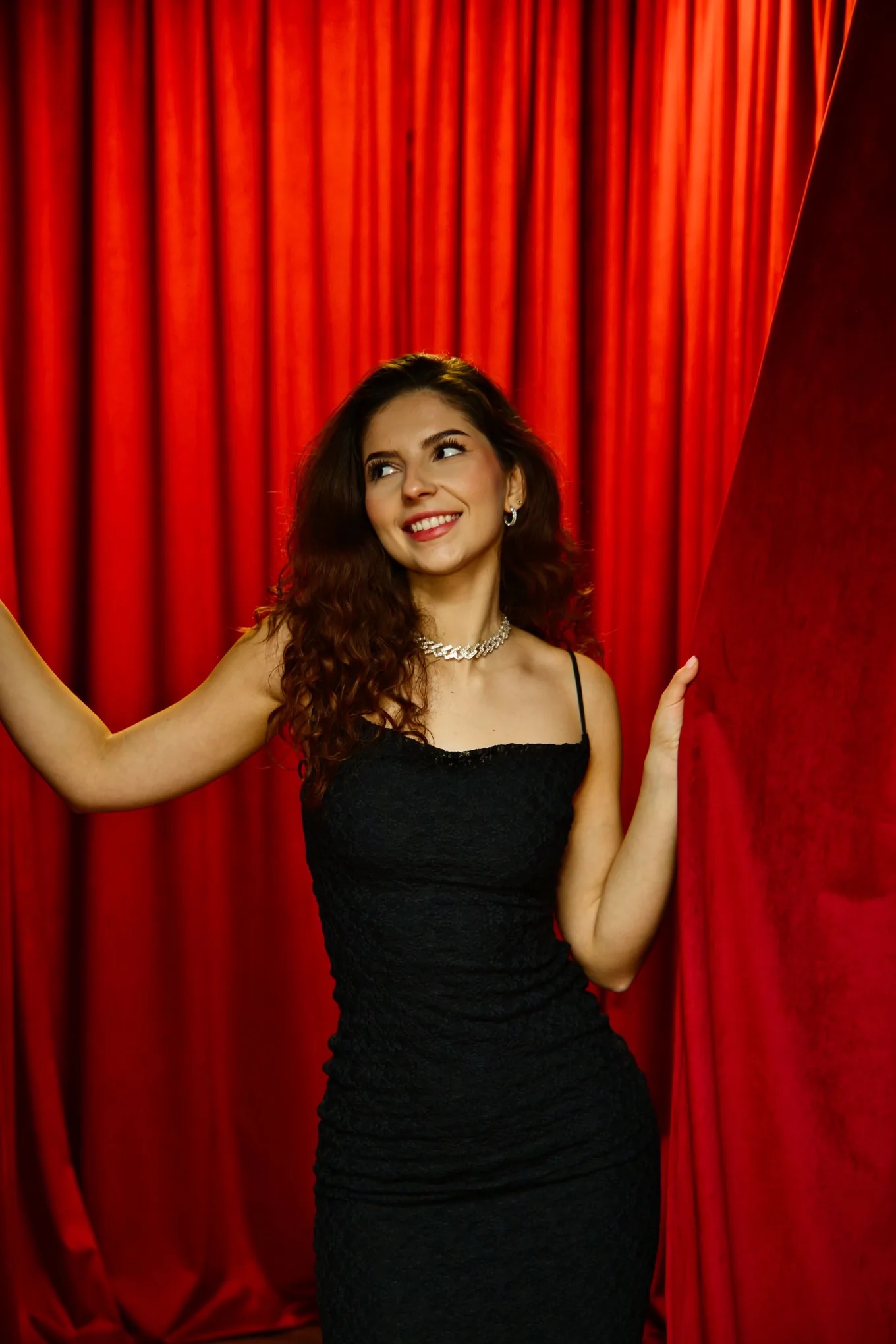Uśmiechnięta brunetka w czarnej sukience i srebrnym naszyjniku, pozująca na tle czerwonej, teatralnej kurtyny. Profesjonalne oświetlenie podkreśla detale stroju.