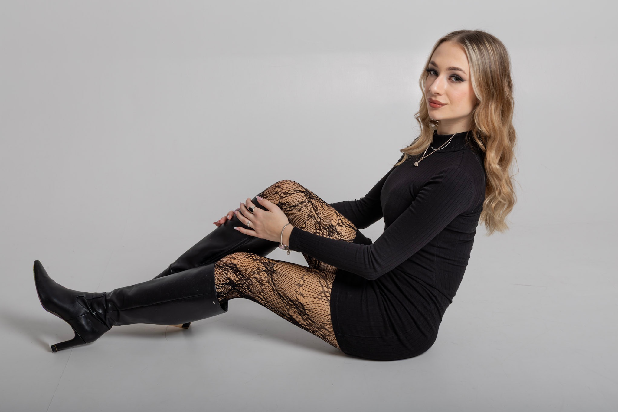 Portret kobiety w czarnej sukience, rajstopach w siatkę i wysokich skórzanych kozakach, siedzącej na szarym tle. Uwagę przykuwa delikatny makijaż i blond włosy.