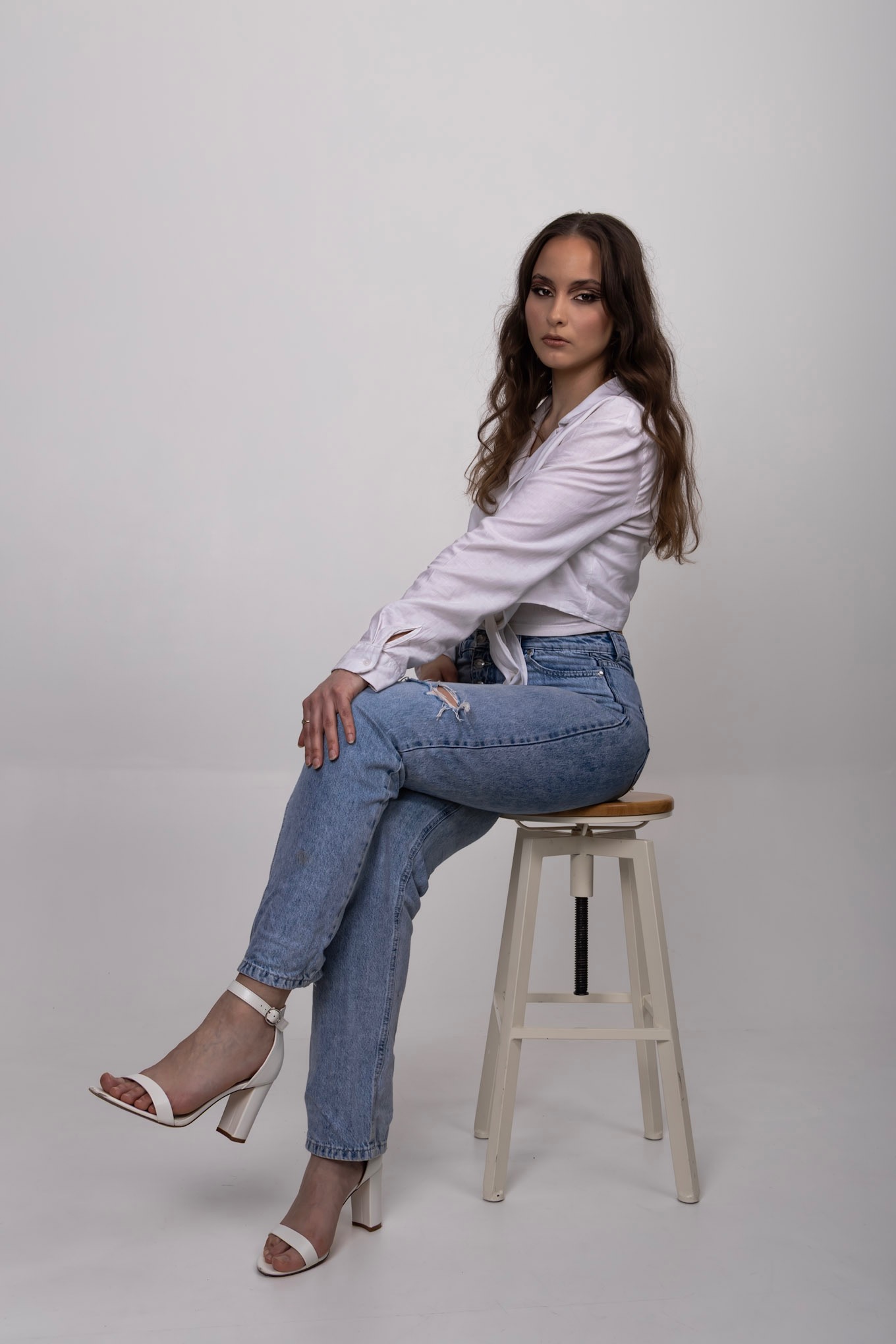 Portret kobiety siedzącej na stołku w białej koszuli i dżinsach z dziurami, na jasnym tle. Uwagę przykuwają białe sandały na obcasie i profesjonalny makijaż.
