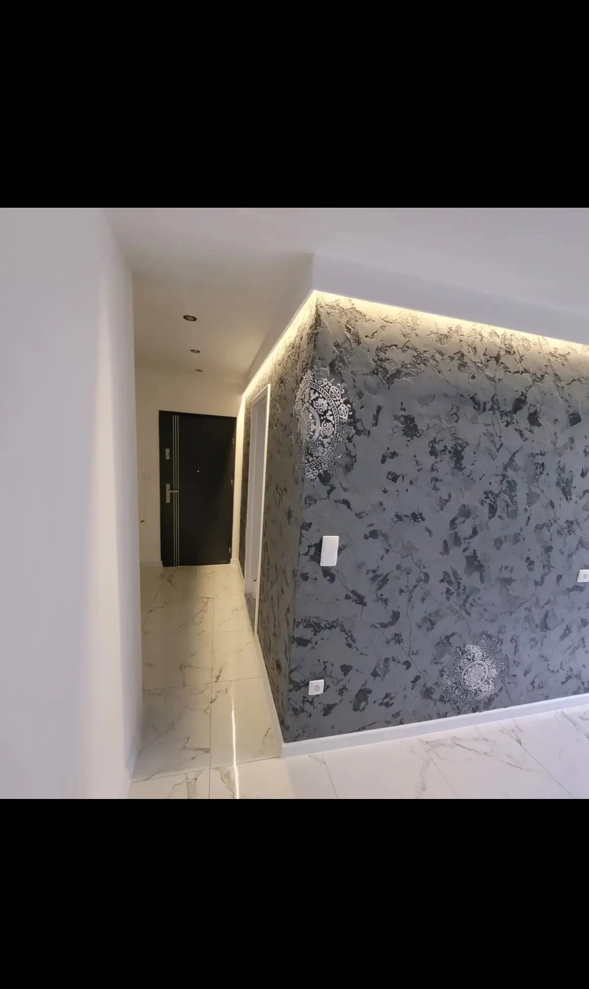 Korytarz z dekoracyjną, szarą ścianą z ornamentami, oświetloną listwą LED. Białe ściany, marmurowe płytki podłogowe i czarne drzwi wejściowe.