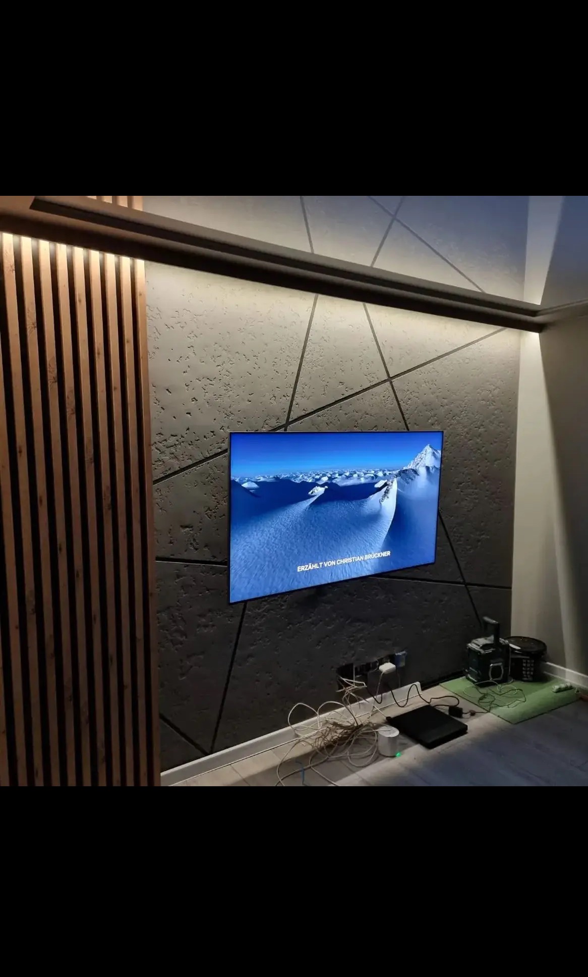 Nowoczesny salon z szarą ścianą o geometrycznym wzorze, drewnianymi lamelami i podświetleniem LED. Telewizor z widokiem górskim. Widoczne kable pod telewizorem.