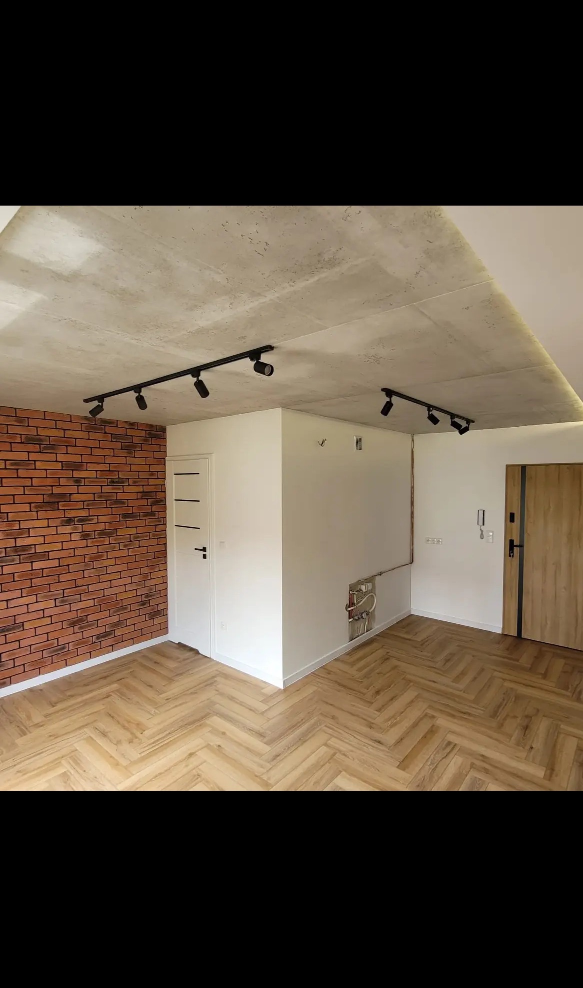 Nowoczesne wnętrze po remoncie: ceglana ściana, parkiet w jodełkę, surowy betonowy sufit z oświetleniem szynowym i białe ściany z instalacją hydrauliczną.