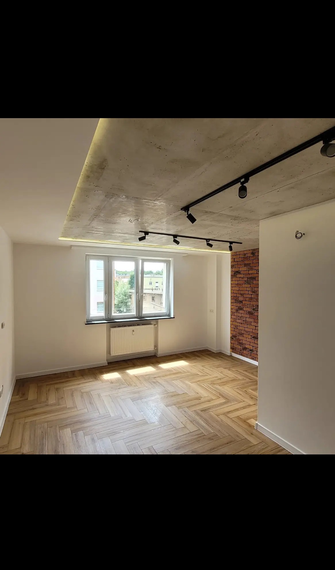 Jasne wnętrze po remoncie: parkiet w jodełkę, białe ściany, sufit betonowy z oświetleniem szynowym i fragment ściany z cegły. Widok na okno z grzejnikiem.
