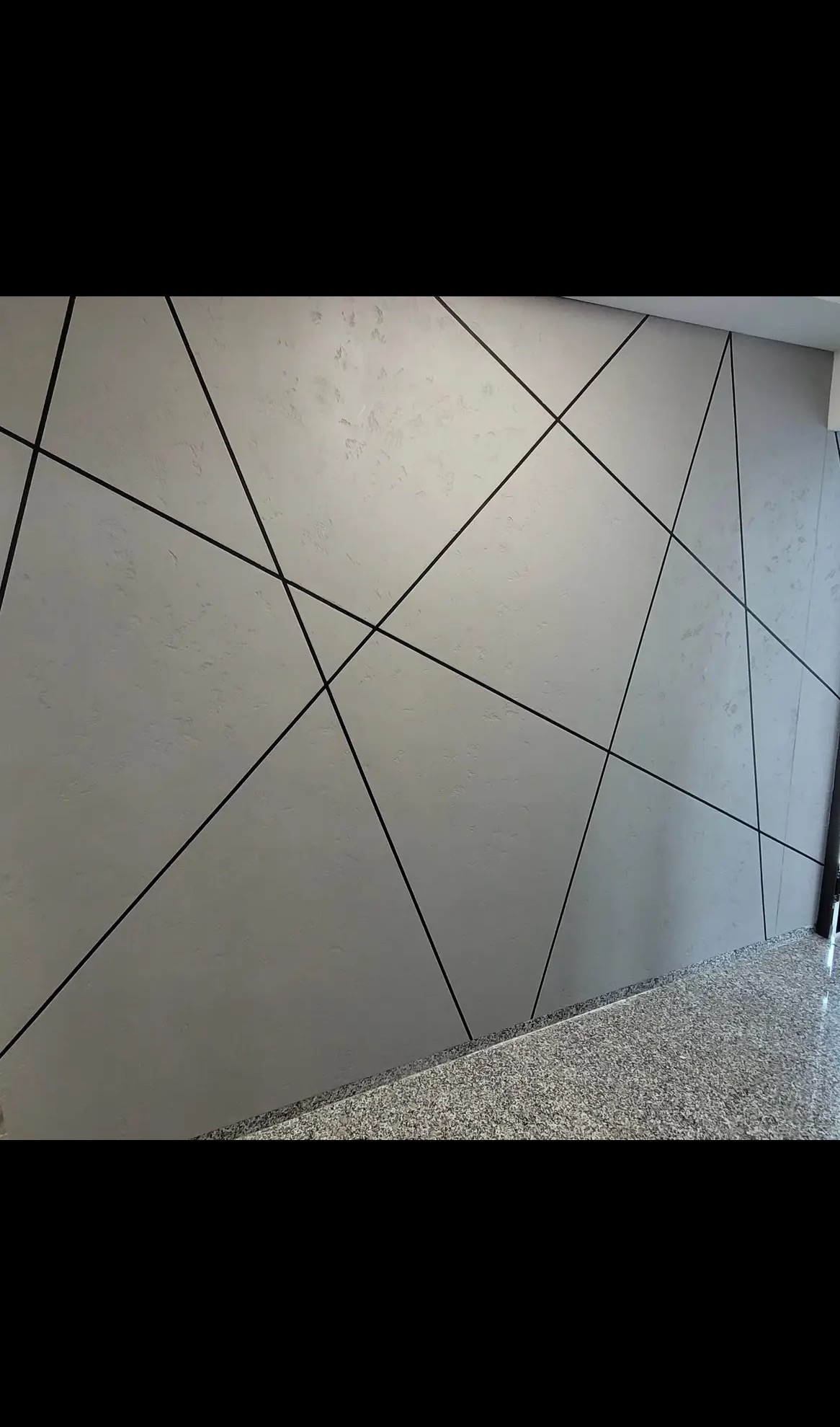 Szara ściana z dekoracyjnymi, czarnymi liniami tworzącymi geometryczny wzór. Widoczna faktura ściany i fragment podłogi z szarego kamienia.