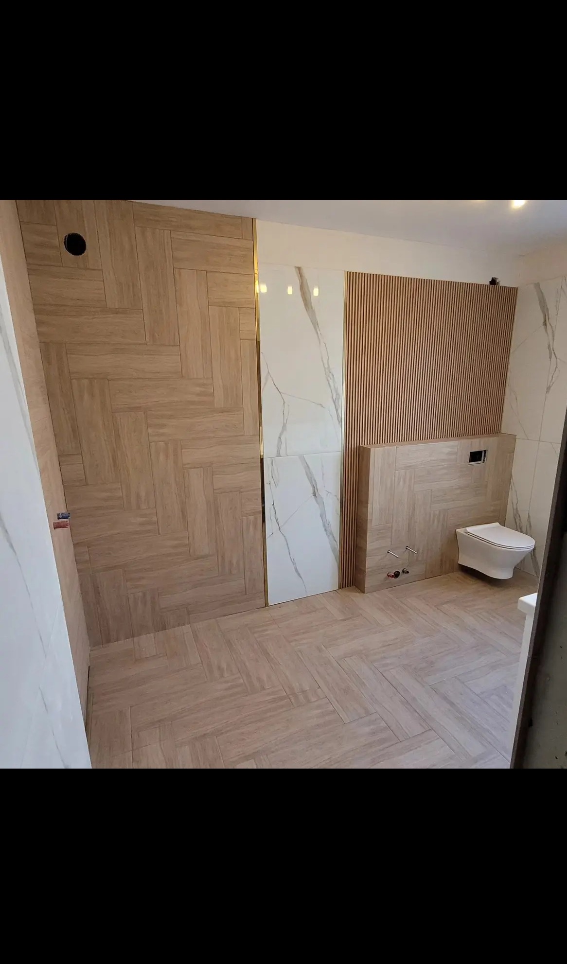 Wykończona łazienka z drewnopodobnymi płytkami ułożonymi w jodełkę na podłodze i ścianach, akcentami z białego marmuru i drewnianymi lamelami. Widoczna toaleta podwieszana.