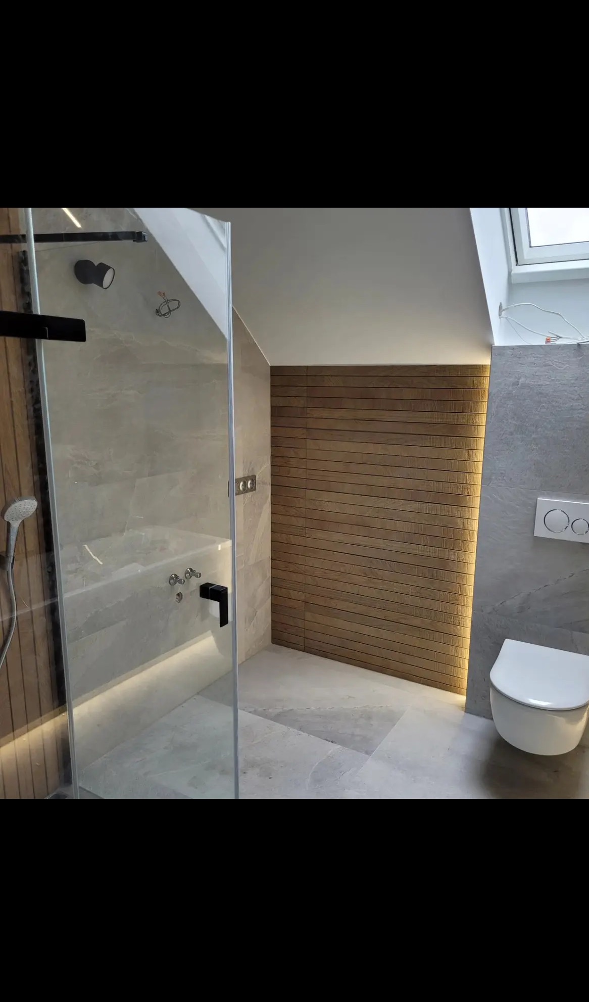 Nowoczesna łazienka z kabiną prysznicową typu walk-in, drewnianą ścianą z podświetleniem LED i wiszącą toaletą. Minimalistyczny design, szare płytki i naturalne materiały.