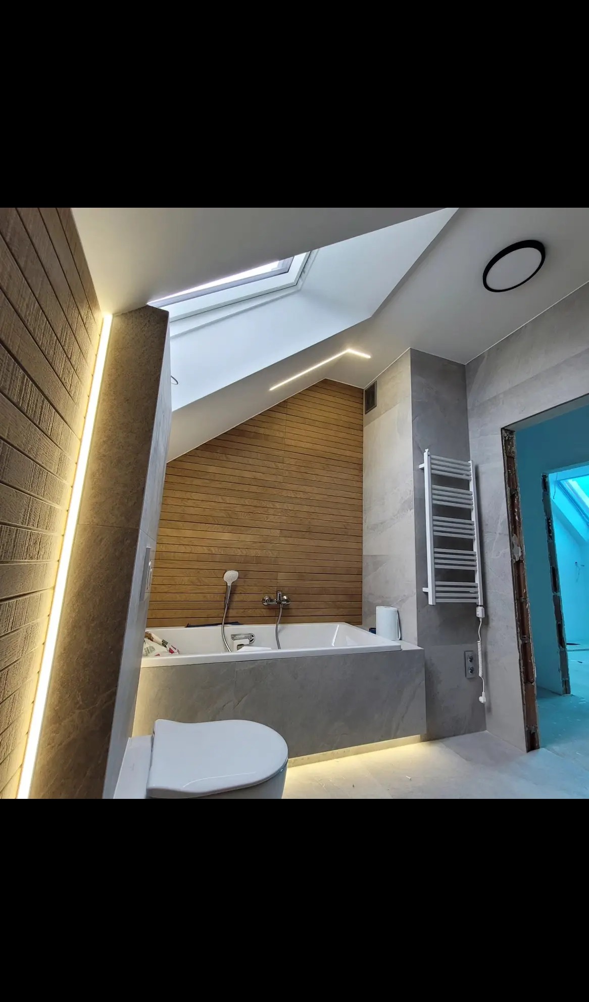 Wykończona łazienka na poddaszu z drewnianą boazerią, wanną obudowaną szarą płytką, grzejnikiem drabinkowym i oświetleniem LED. Widoczne okno dachowe.