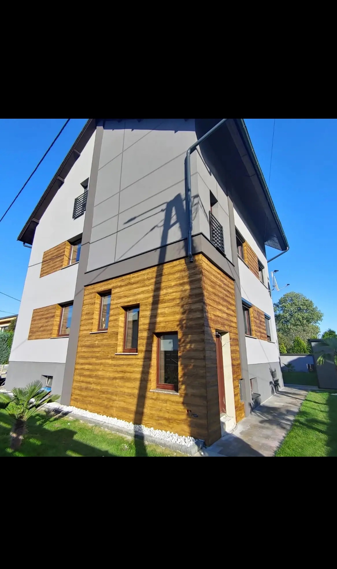 Elewacja domu z szarymi panelami i drewnopodobną okładziną, widok z dołu w słoneczny dzień. Kontrastowe połączenie materiałów i nowoczesna architektura.