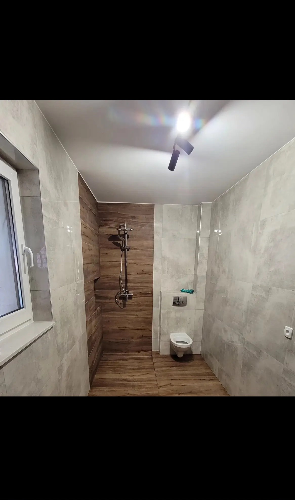 Wykończona łazienka z drewnopodobnymi i szarymi płytkami, kabiną prysznicową i toaletą. Nowoczesne oświetlenie sufitowe. Widok na okno z białymi ramami.