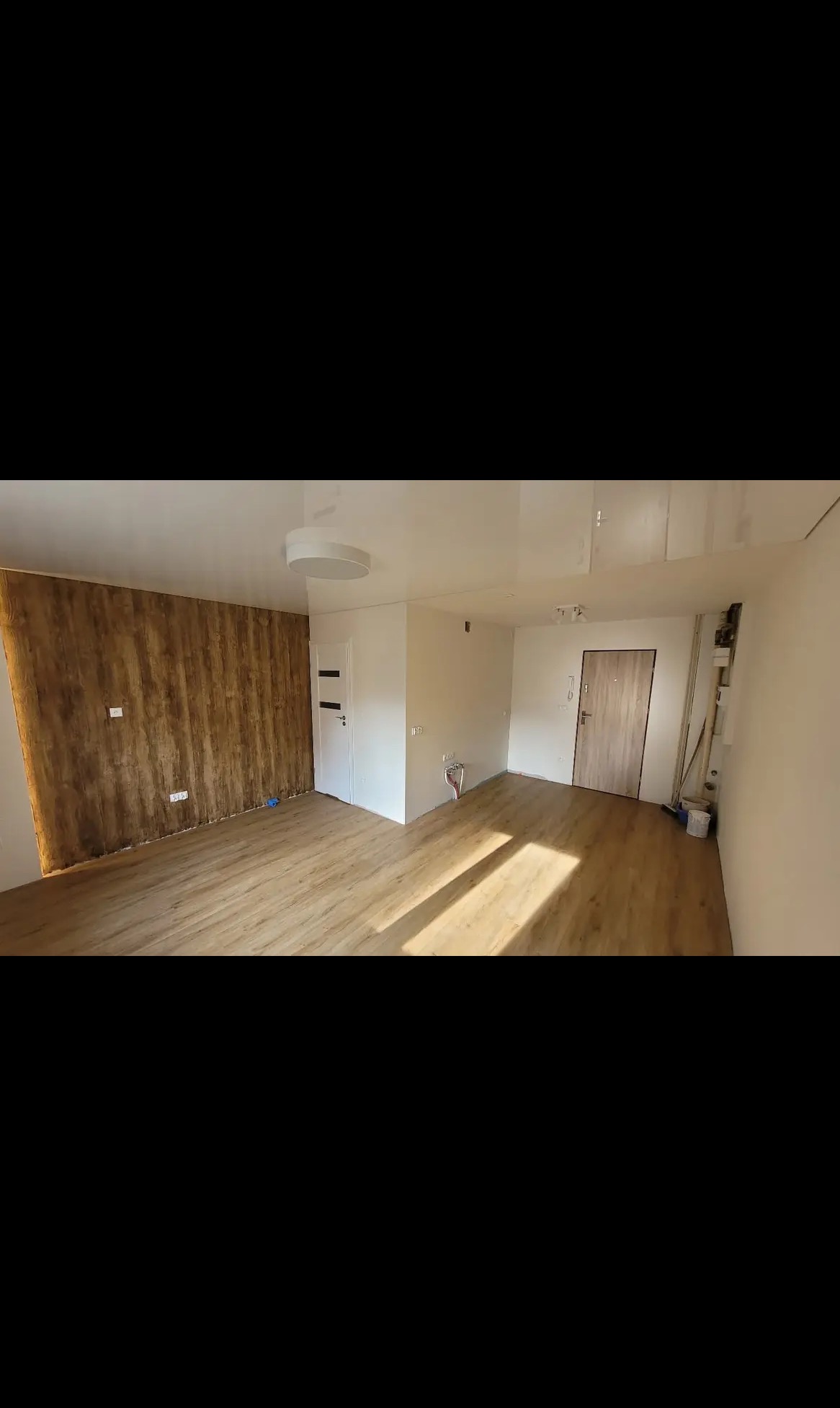 Jasne wnętrze po remoncie: drewniana podłoga, ściana z efektem drewna, biały sufit napinany i drzwi. Widoczne instalacje elektryczne.