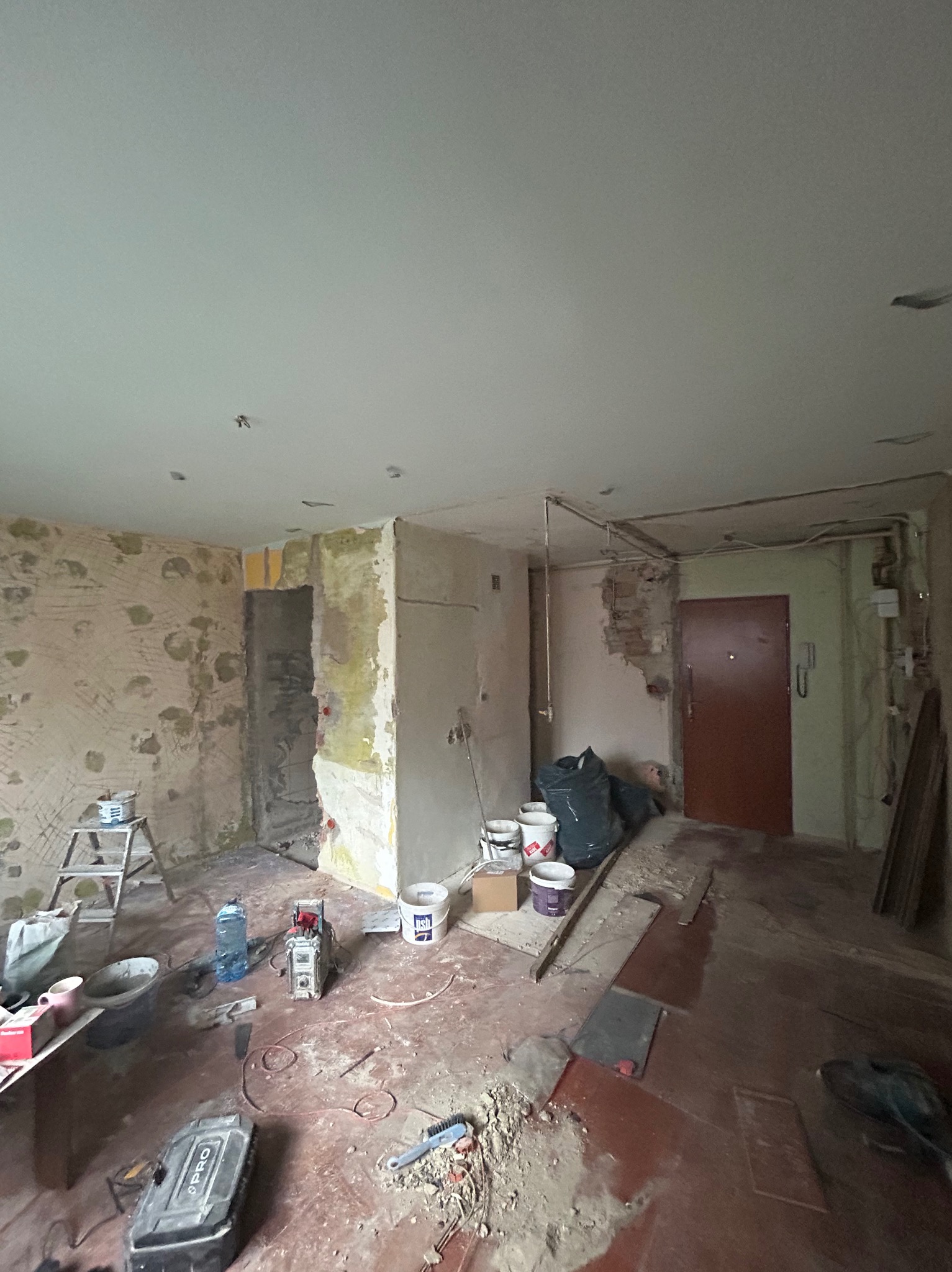 Wnętrze w trakcie remontu: ściany przygotowane do malowania, widoczne narzędzia, worki z gruzem i puste wiadra po farbach. Podłoga częściowo zakryta.