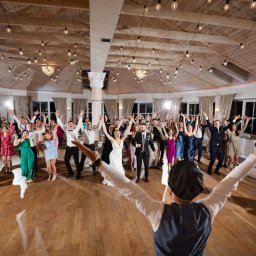 DJ Oskar Łakomy - Rozbawieni goście weselni z podniesionymi rękami, prowadzeni przez wodzireja w kapeluszu, w przestronnej sali z drewnianym sufitem i dekoracyjnym oświetleniem.