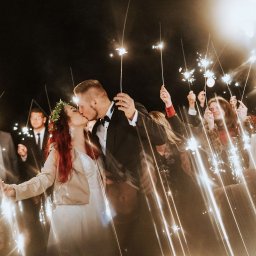 DJ Oskar Łakomy - Pocałunek nowożeńców otoczonych gośćmi z zimnymi ogniami, nocne wesele z iskrzącymi się światłami, panna młoda w wianku, pan młody w smokingu.