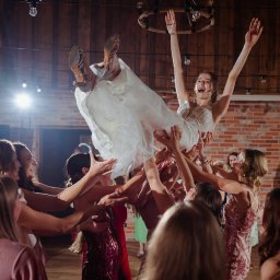 DJ Oskar Łakomy - Radosna panna młoda w białej sukni, uniesiona w górę przez grupę kobiet podczas wesela w rustykalnej sali z ceglaną ścianą i drewnianym parkietem.