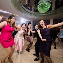 DJ Oskar Łakomy - Rozbawione kobiety w sukienkach tańczą na parkiecie pod kryształowym żyrandolem podczas imprezy. W tle widoczne oświetlenie sceniczne i sylwetki muzyków.