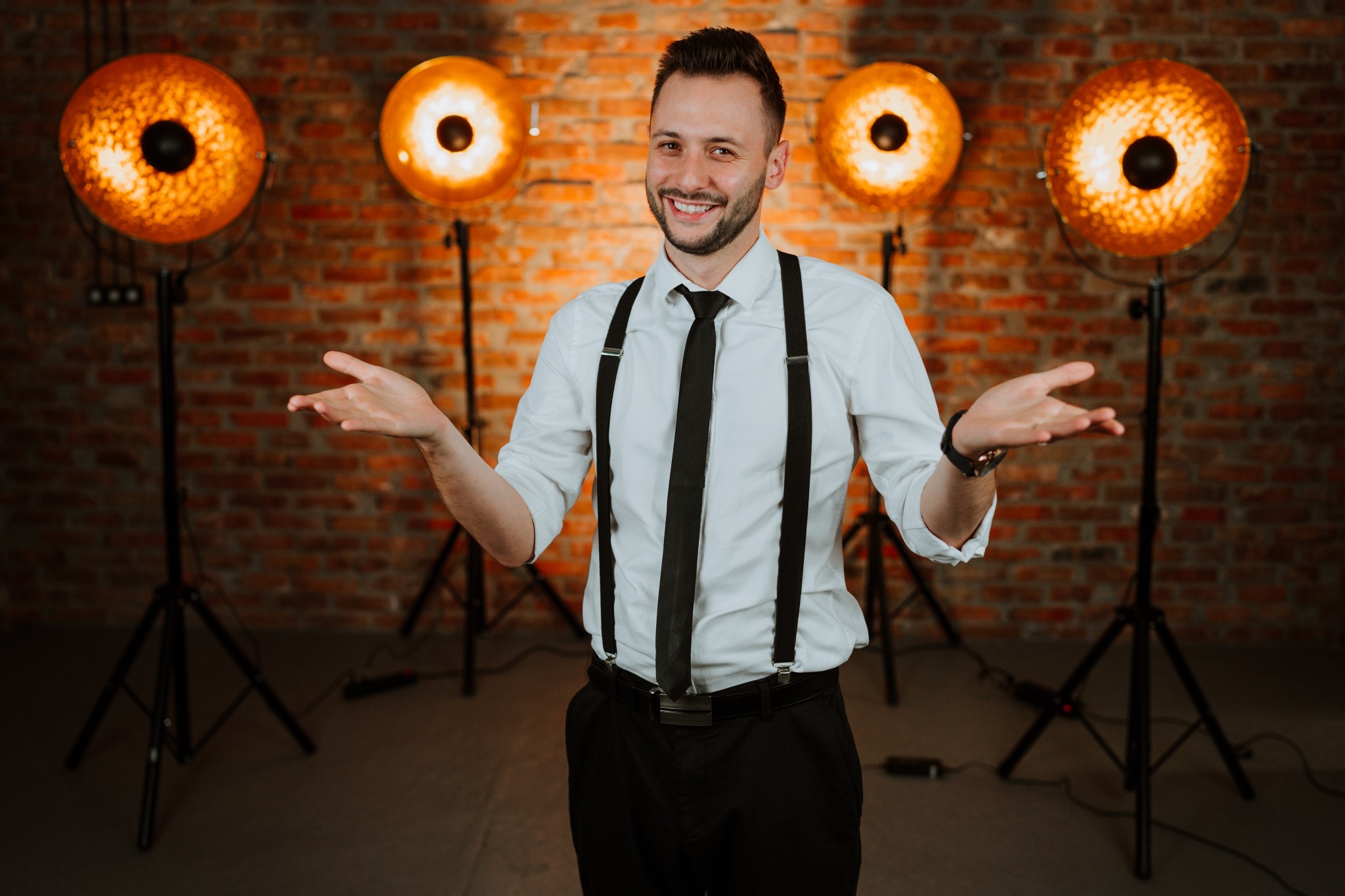 Uśmiechnięty mężczyzna w szelkach i krawacie, na tle ceglanej ściany z czterema lampami studyjnymi, prezentuje ofertę z otwartymi dłońmi.