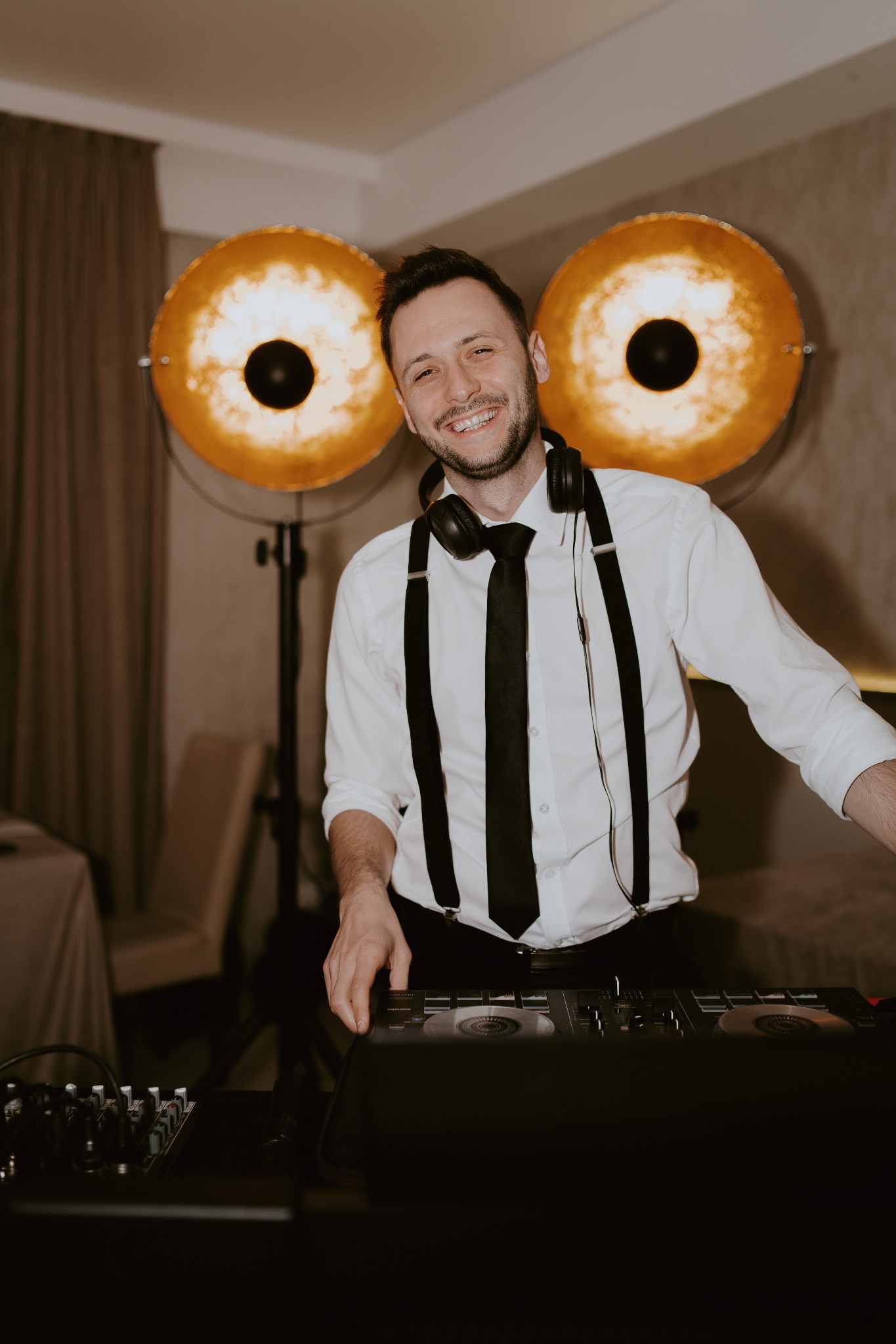 Uśmiechnięty DJ w białej koszuli z szelkami i słuchawkami na szyi, obsługujący sprzęt DJ-ski. W tle okrągłe lampy o ciepłym świetle.