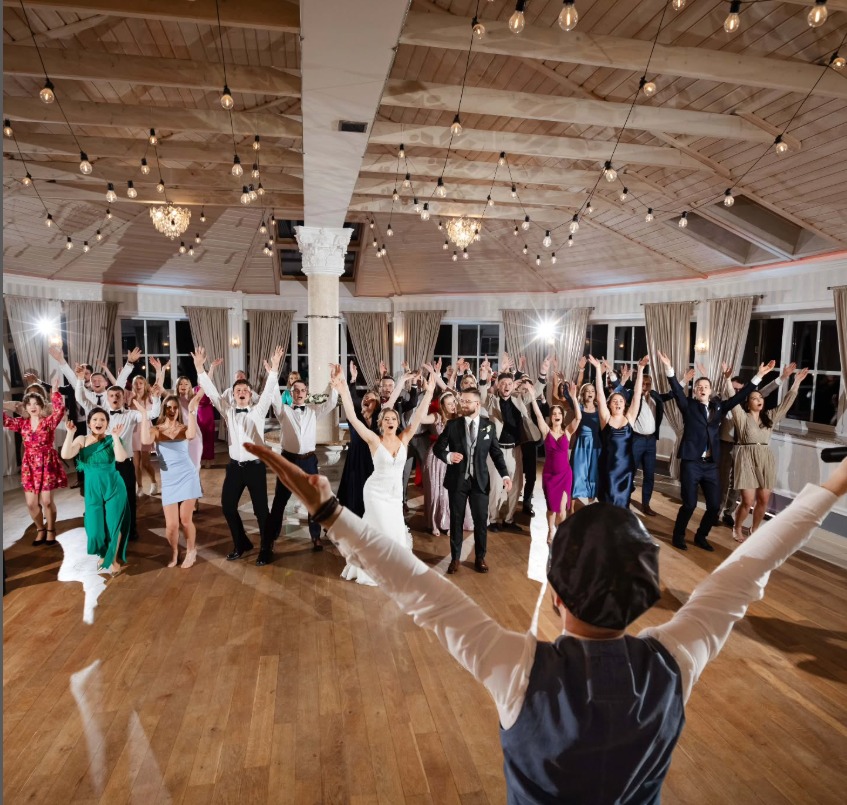 Rozbawieni goście weselni z podniesionymi rękami, prowadzeni przez wodzireja w kapeluszu, w przestronnej sali z drewnianym sufitem i dekoracyjnym oświetleniem.