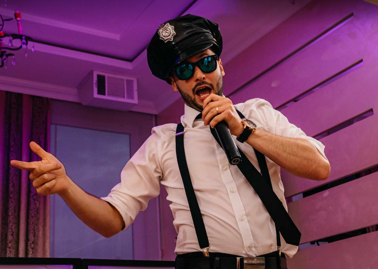 Mężczyzna w stroju policjanta śpiewa do mikrofonu, gestykulując. Ubrany w białą koszulę, szelki i czapkę. Scena oświetlona fioletowym światłem.