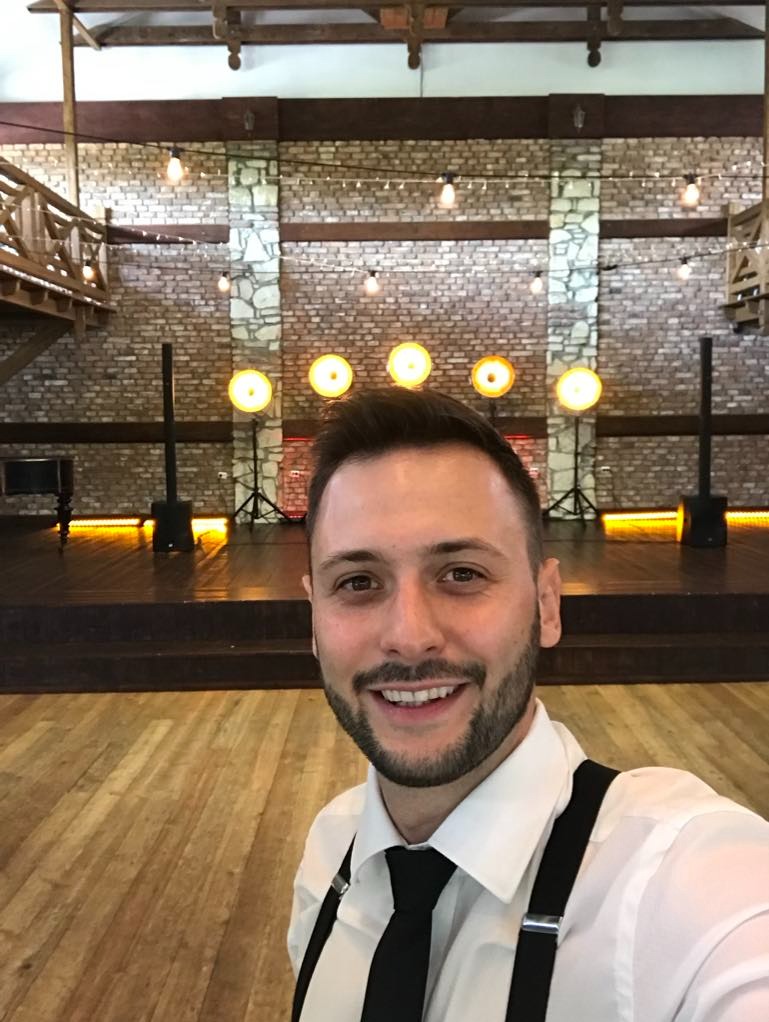 Uśmiechnięty mężczyzna w koszuli i krawacie na tle sceny z oświetleniem punktowym i nagłośnieniem. Widoczna ceglana ściana i drewniana podłoga.