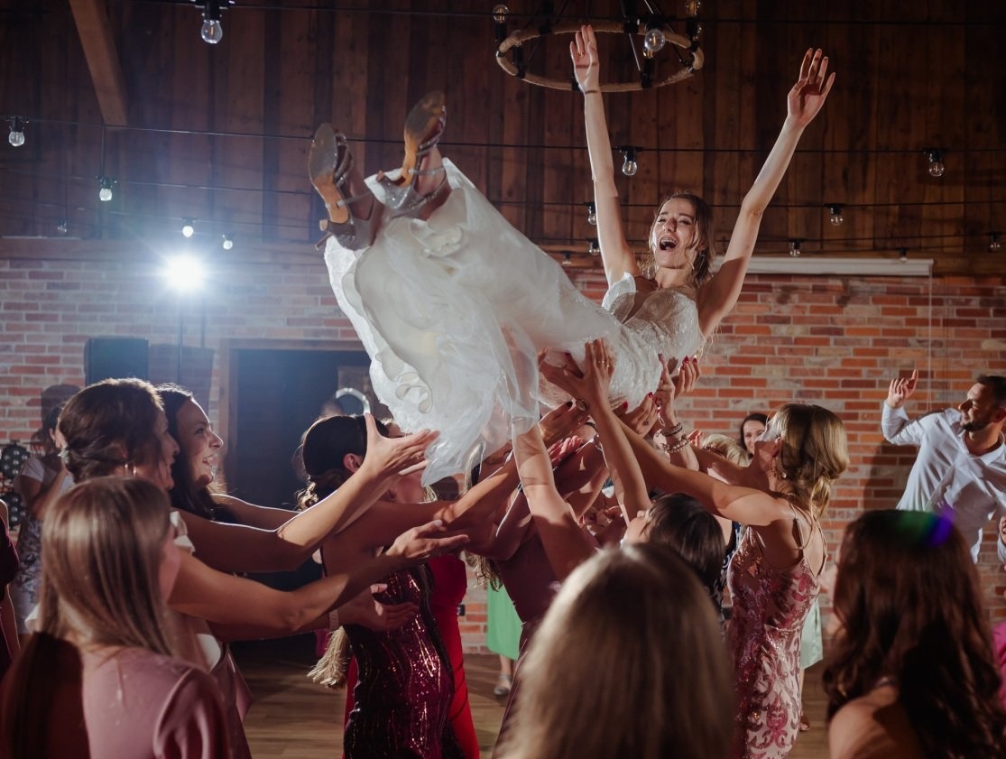 Radosna panna młoda w białej sukni, uniesiona w górę przez grupę kobiet podczas wesela w rustykalnej sali z ceglaną ścianą i drewnianym parkietem.