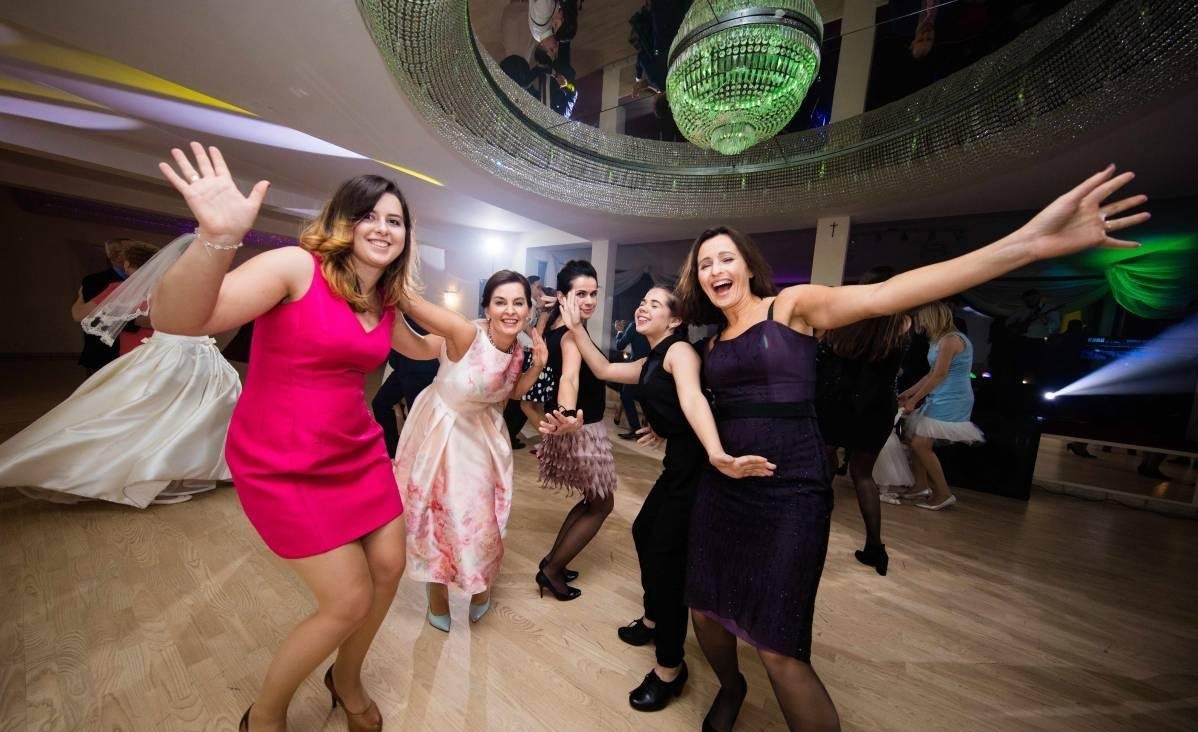 Rozbawione kobiety w sukienkach tańczą na parkiecie pod kryształowym żyrandolem podczas imprezy. W tle widoczne oświetlenie sceniczne i sylwetki muzyków.