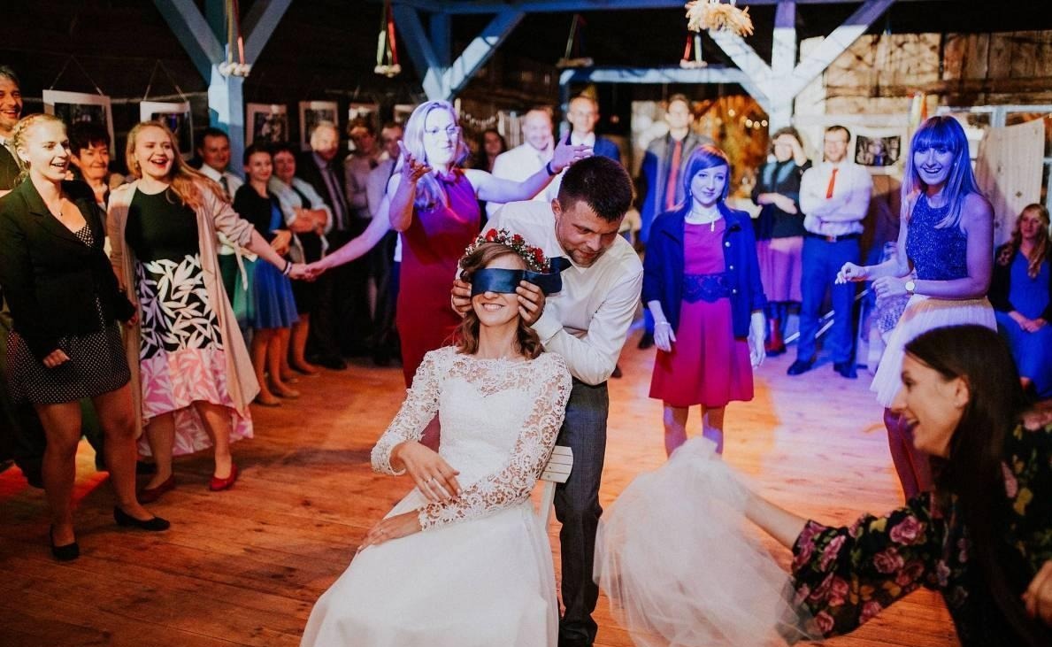 Zabawa weselna: Pan młody zawiązuje oczy siedzącej pannie młodej, wokół goście weselni na drewnianym parkiecie. Dekoracje rustykalne, ciepłe oświetlenie.