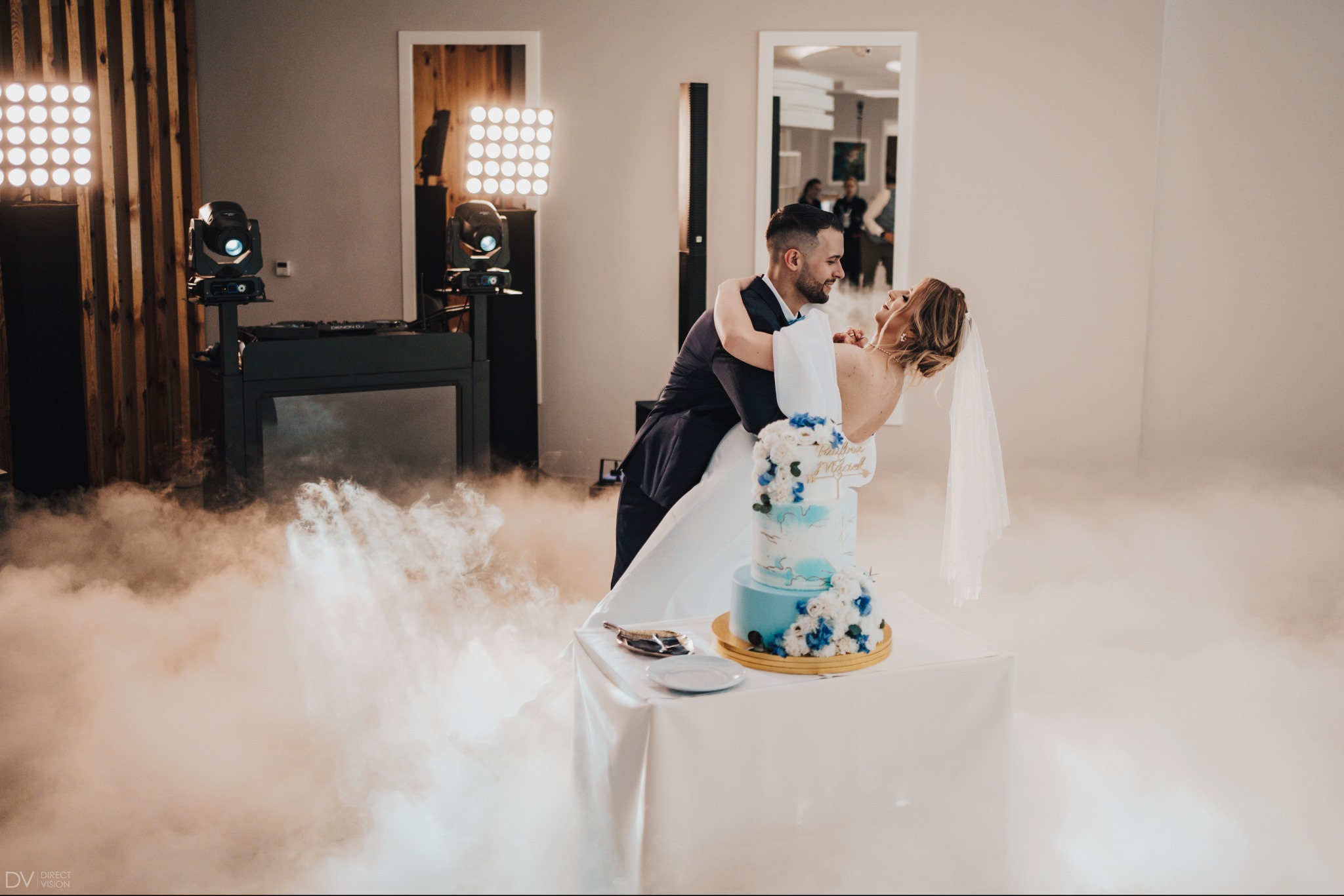 Para młoda w tańcu, tort weselny na pierwszym planie, dekoracje świetlne i dym tworzą romantyczny nastrój. DJ-ka w tle. Ujęcie z dołu.