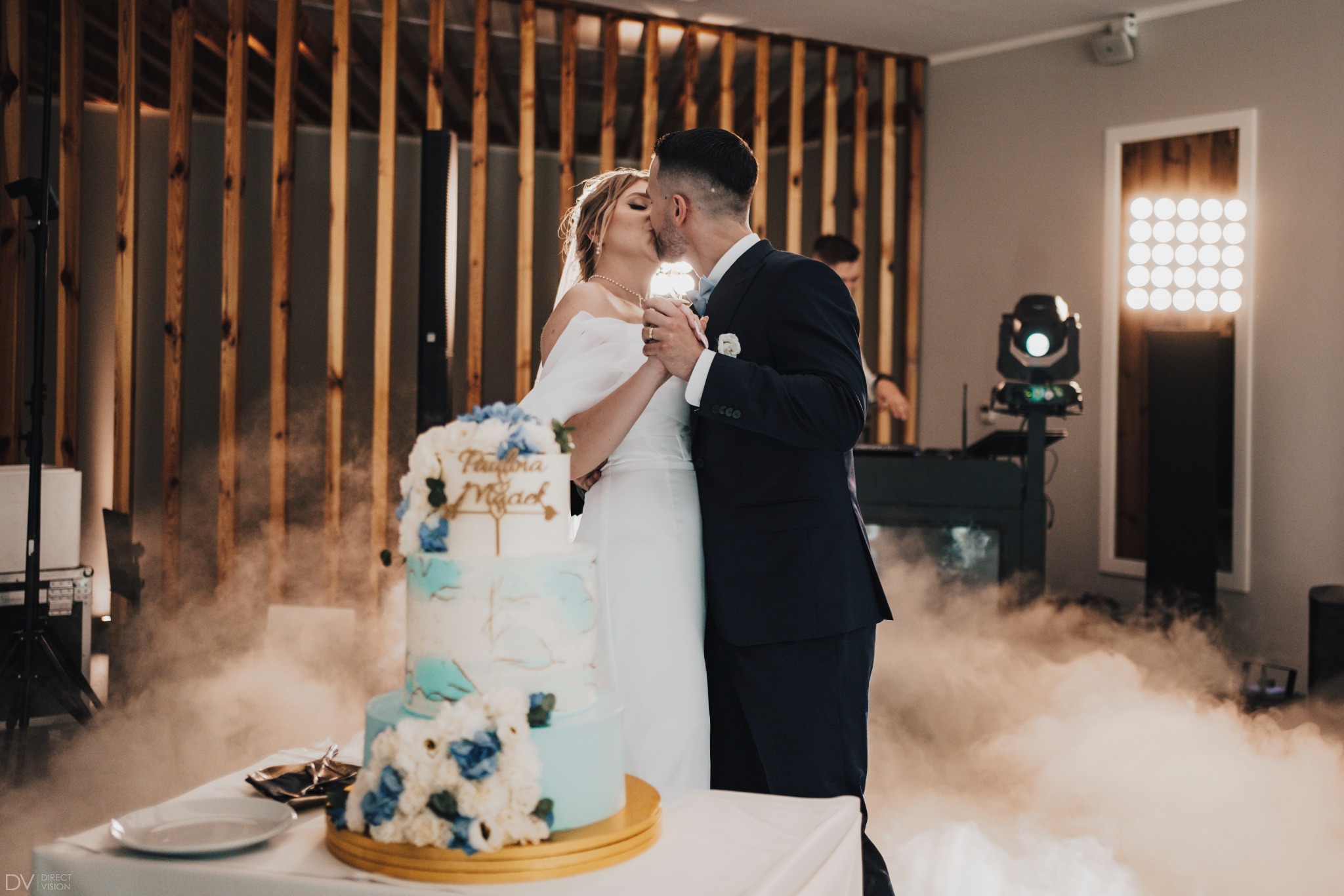 Pocałunek pary młodej za tortem weselnym w dymie scenicznym, w tle drewniana ścianka i oświetlenie. Ujęcie z bliska, ciepłe światło.