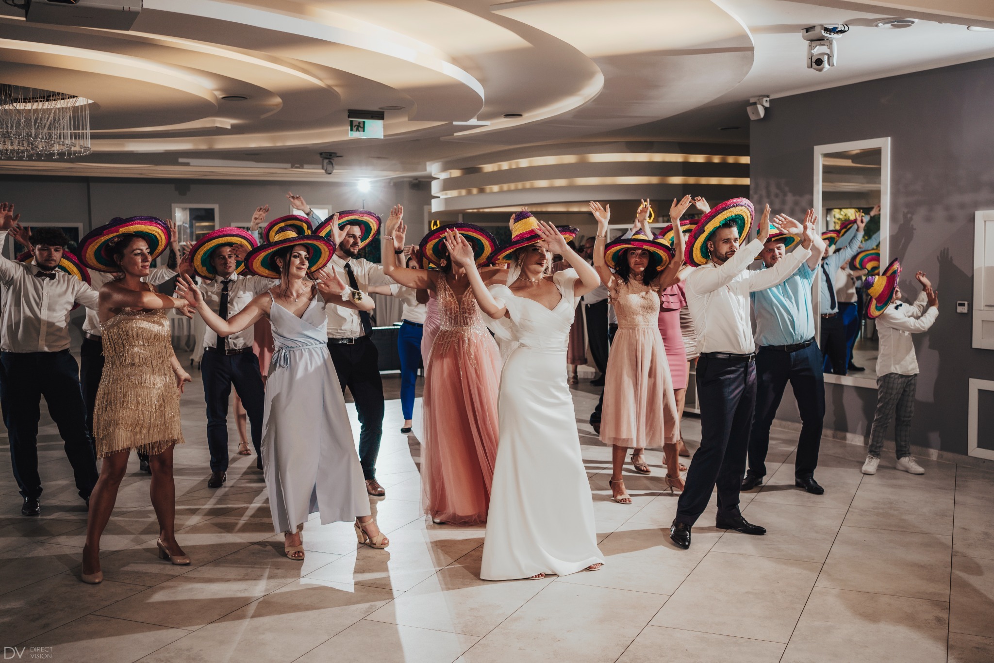 Roztańczona grupa gości weselnych w kolorowych sombrerach, w eleganckiej sali balowej z parkietem w jasne kwadraty i dekoracyjnym oświetleniem sufitowym.