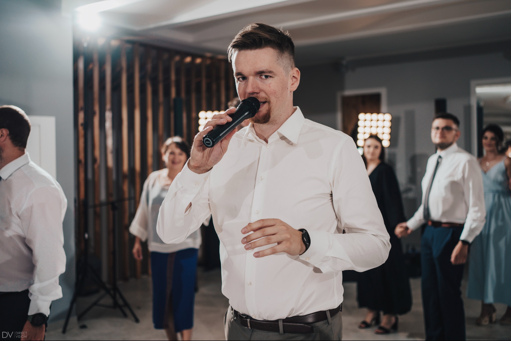 Mężczyzna z mikrofonem w białej koszuli, otoczony przez gości weselnych. W tle drewniana ściana i oświetlenie. Ujęcie z perspektywy widza, skupione na wokaliście.