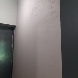 Uliana Boitsun - Ściana z dekoracyjną, szarą strukturą obok ciemnej ściany i fragmentu ościeżnicy drzwi. Tekstura ściany z bliska, widoczne nierówności i wzór.