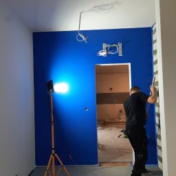 Uliana Boitsun - Ściana w intensywnym odcieniu niebieskiego, oświetlona lampą budowlaną, malowana przez pracownika. Widoczne przygotowania do prac wykończeniowych.