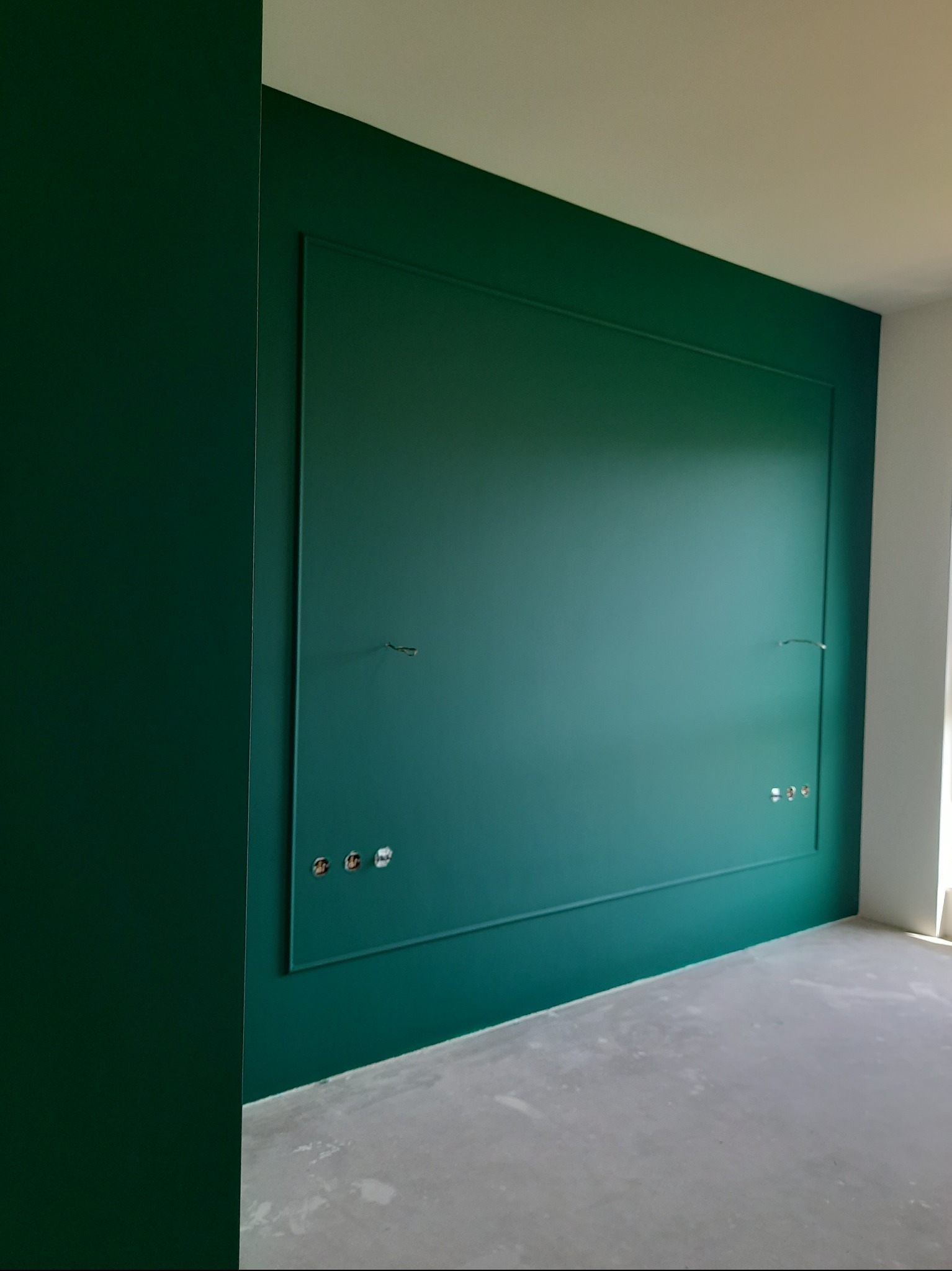 Szmaragdowa ściana z dekoracyjną ramą i gniazdkami elektrycznymi w minimalistycznym wnętrzu, świeżo po malowaniu. Widoczne niedoskonałości podłoża.