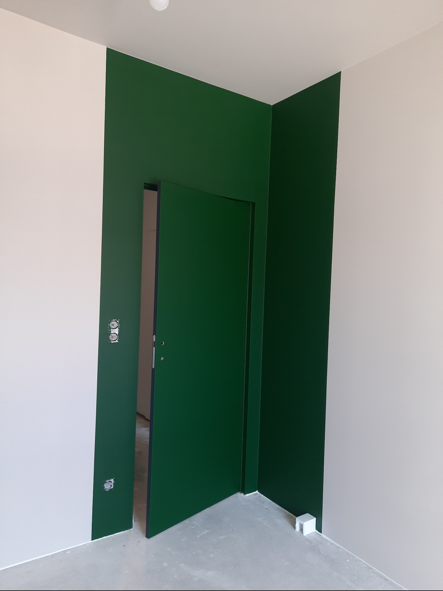 Ściana pomalowana na zielono z otwartymi drzwiami w tym samym kolorze, kontrastująca z białą ścianą. Widoczne gniazdka elektryczne i surowa podłoga.