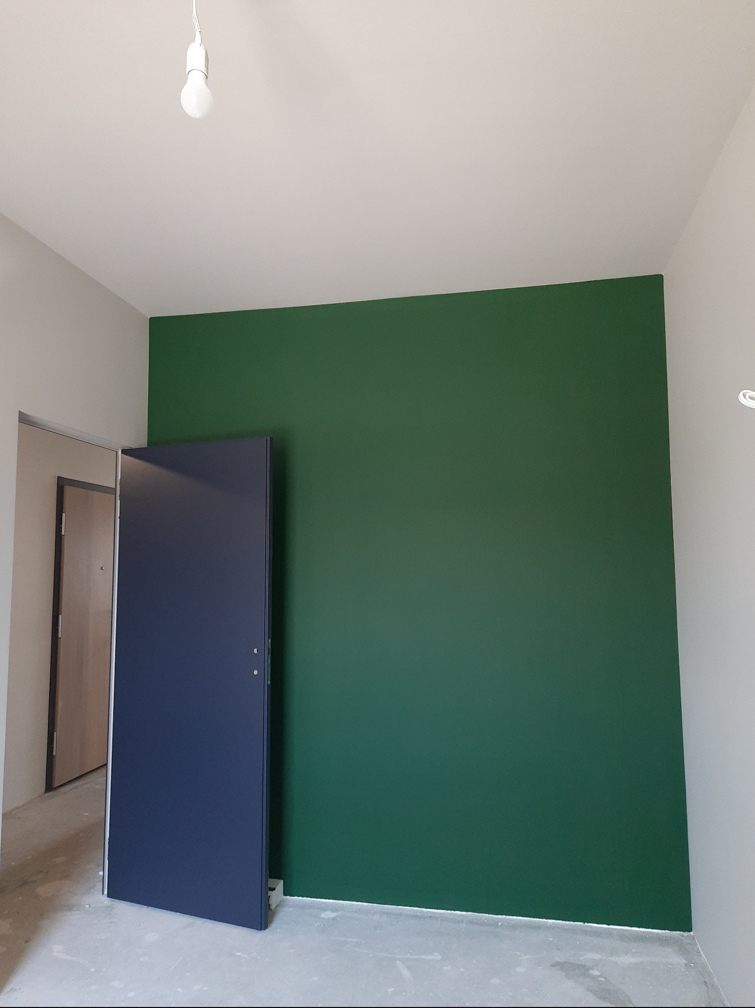 Ściana pomalowana na zielono w nowym mieszkaniu, z niebieskimi drzwiami i surową podłogą. Widoczna żarówka i fragment ściany w kolorze szarym.