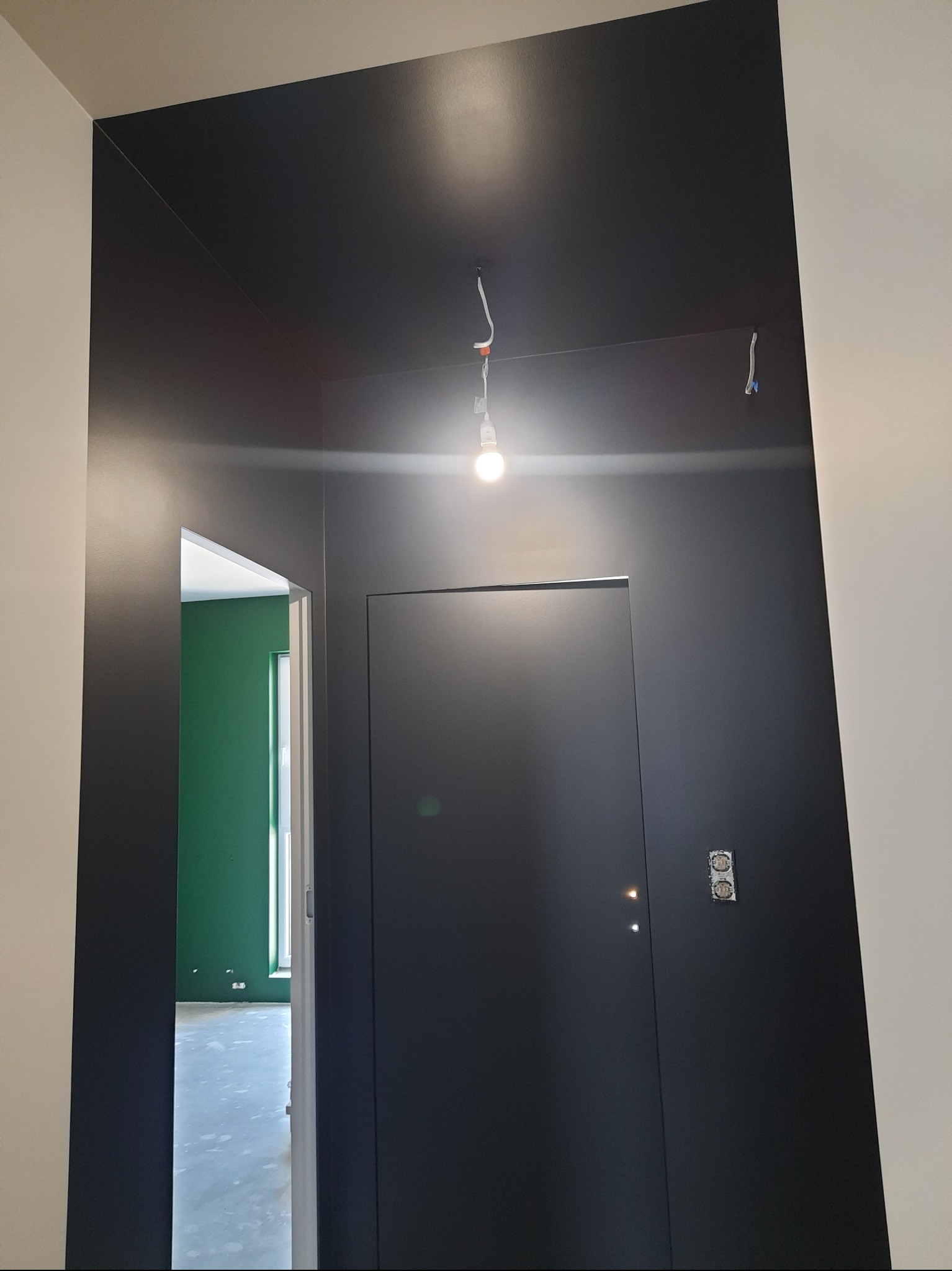 Ciemna ściana z drzwiami i wystającym kablem żarówki, widok na zielony pokój. Gładka powierzchnia, świeżo po pracach wykończeniowych, z widocznymi gniazdkami elektrycznymi.