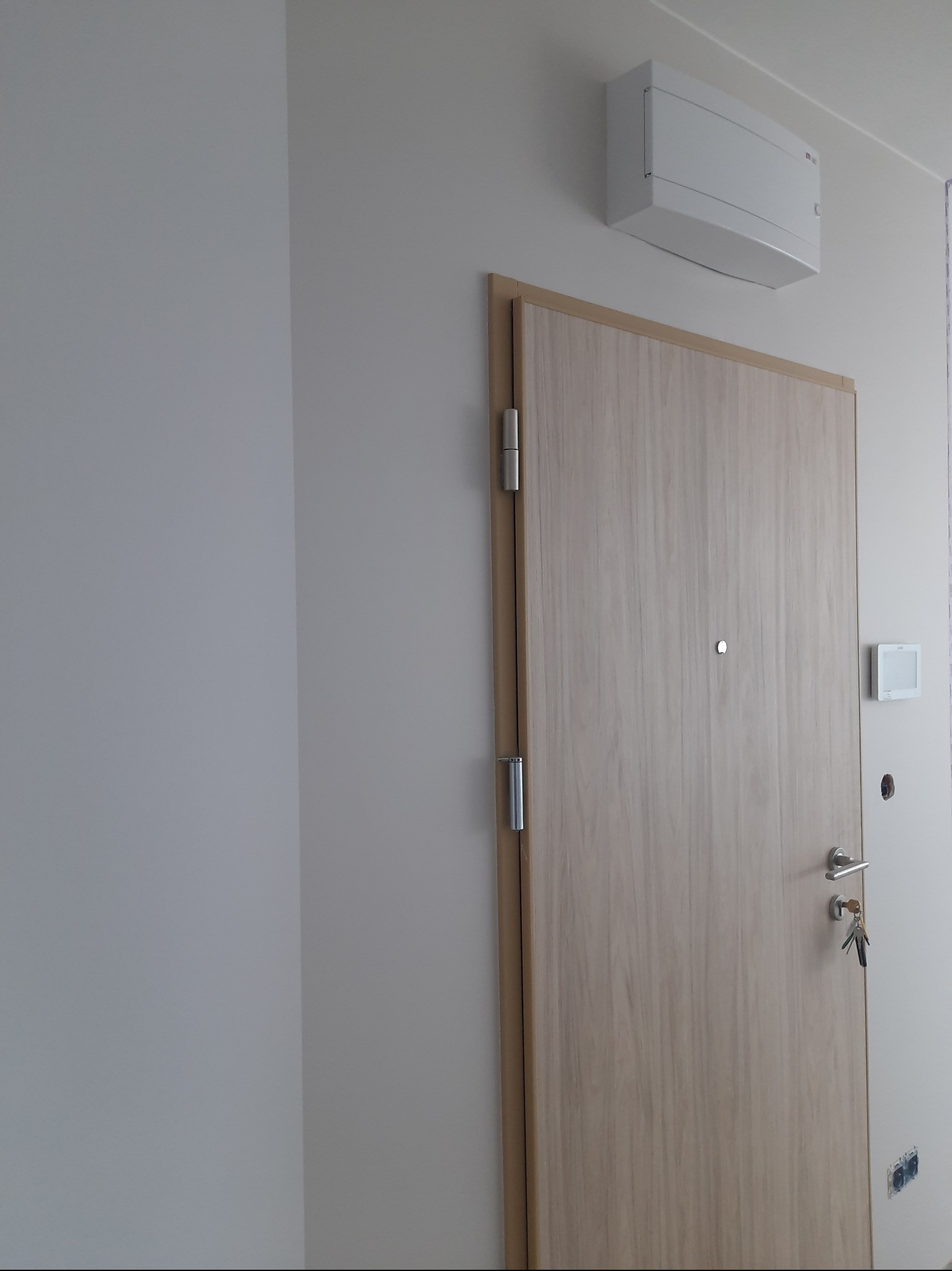 Jasne, świeżo pomalowane ściany w przedpokoju z drewnianymi drzwiami wejściowymi i klimatyzatorem na ścianie. Widoczne gniazdka elektryczne.