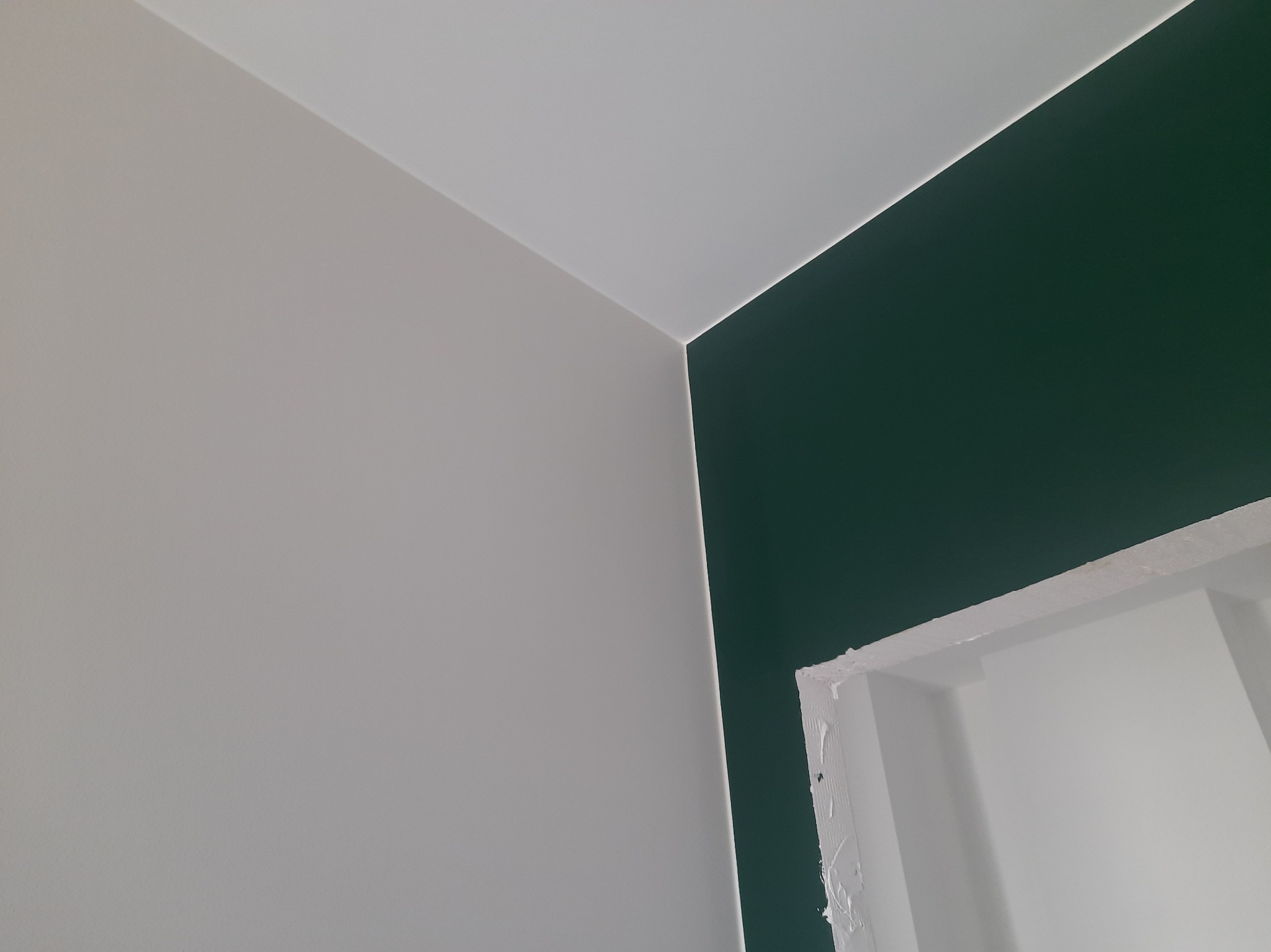 Ściana pomalowana na dwa kolory: szary i butelkowa zieleń. Widoczny fragment ościeżnicy. Minimalistyczne ujęcie detalu wykończenia wnętrza.