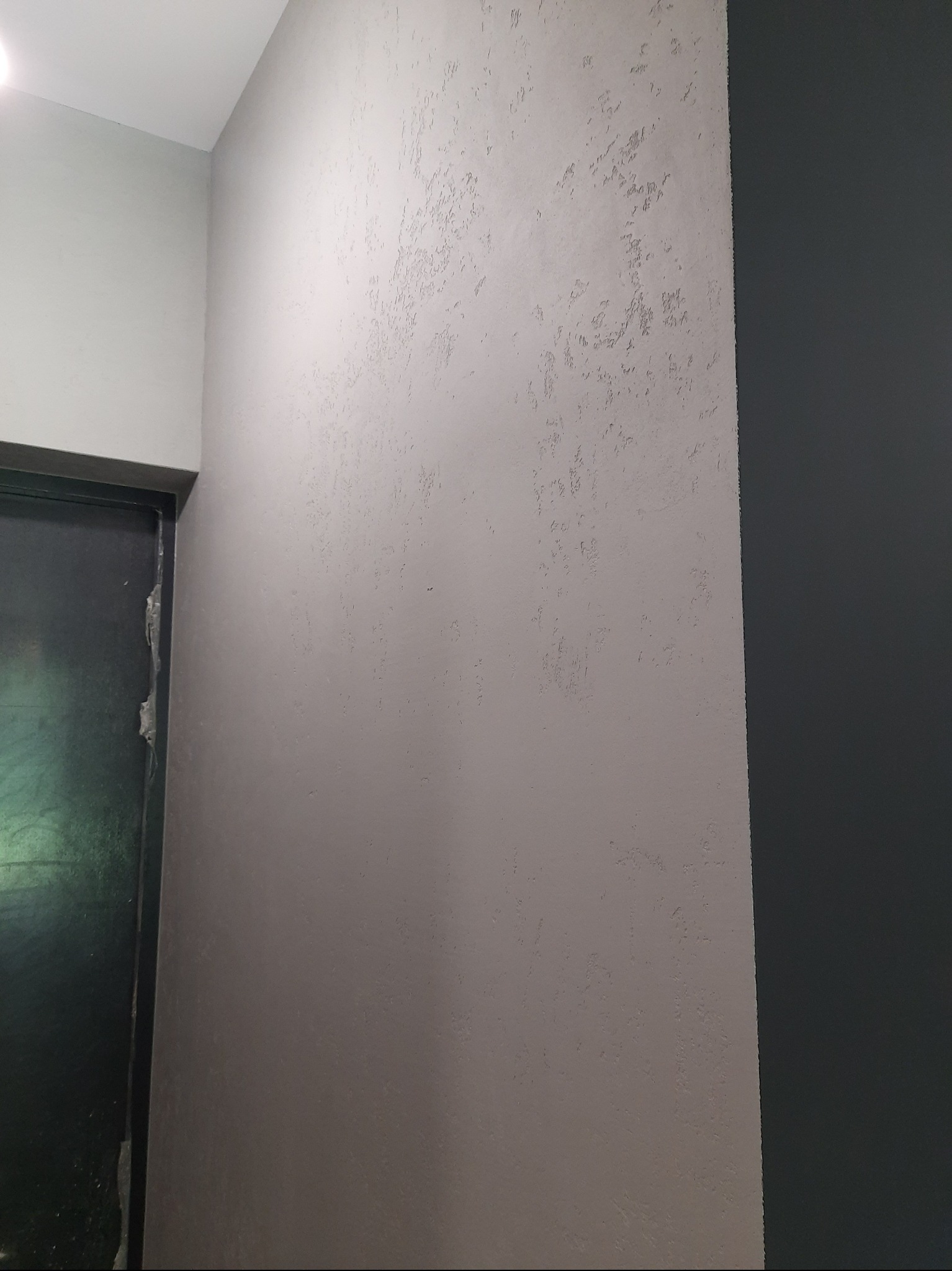 Ściana z dekoracyjną, szarą strukturą obok ciemnej ściany i fragmentu ościeżnicy drzwi. Tekstura ściany z bliska, widoczne nierówności i wzór.