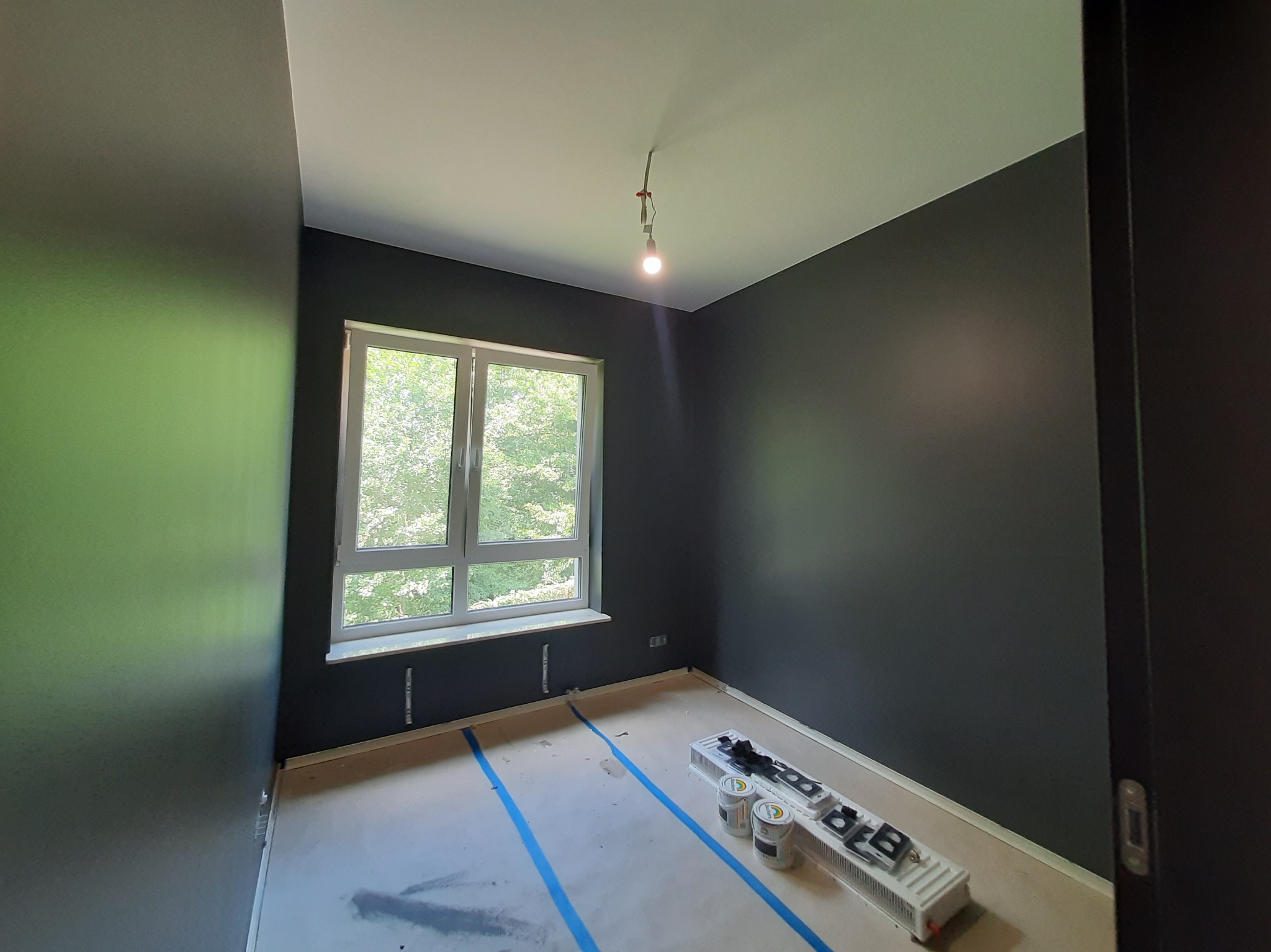 Pokój po malowaniu na ciemny kolor, widoczne puszki z farbą i wałek malarski. Podłoga zabezpieczona folią i taśmą malarską. Okno z widokiem na zieleń.