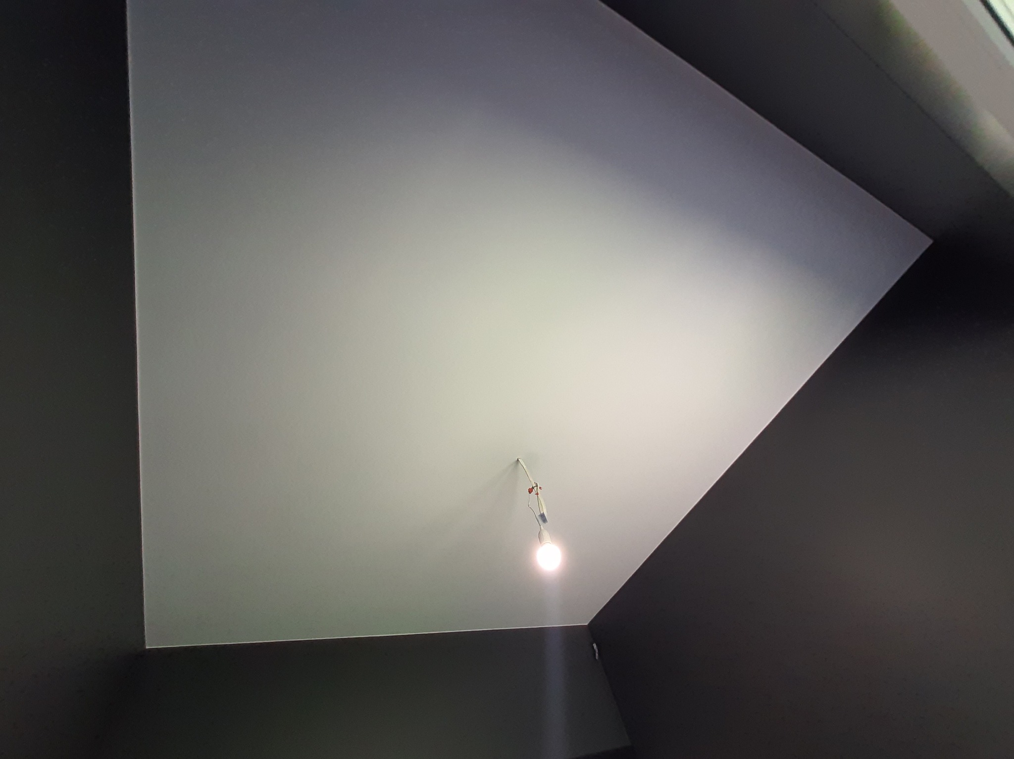 Gładki, pomalowany sufit w odcieniach szarości z wiszącą żarówką na kablu. Ściany pomalowane na ciemny kolor. Ujęcie z dołu, podkreślające geometrię pomieszczenia.