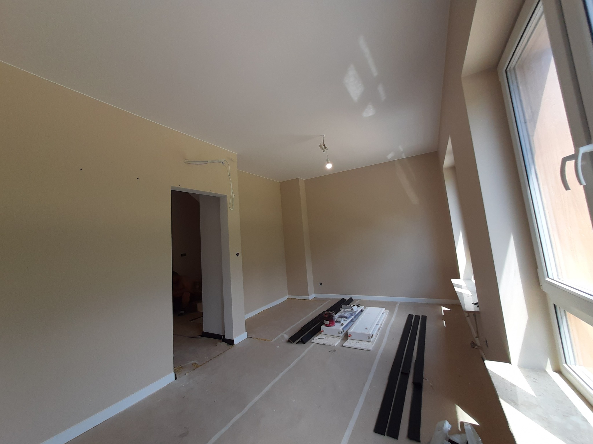 Jasne wnętrze po malowaniu ścian na beżowo, z listwami przypodłogowymi i elementami do montażu paneli pod oknem. Widoczne przygotowania do układania podłogi.