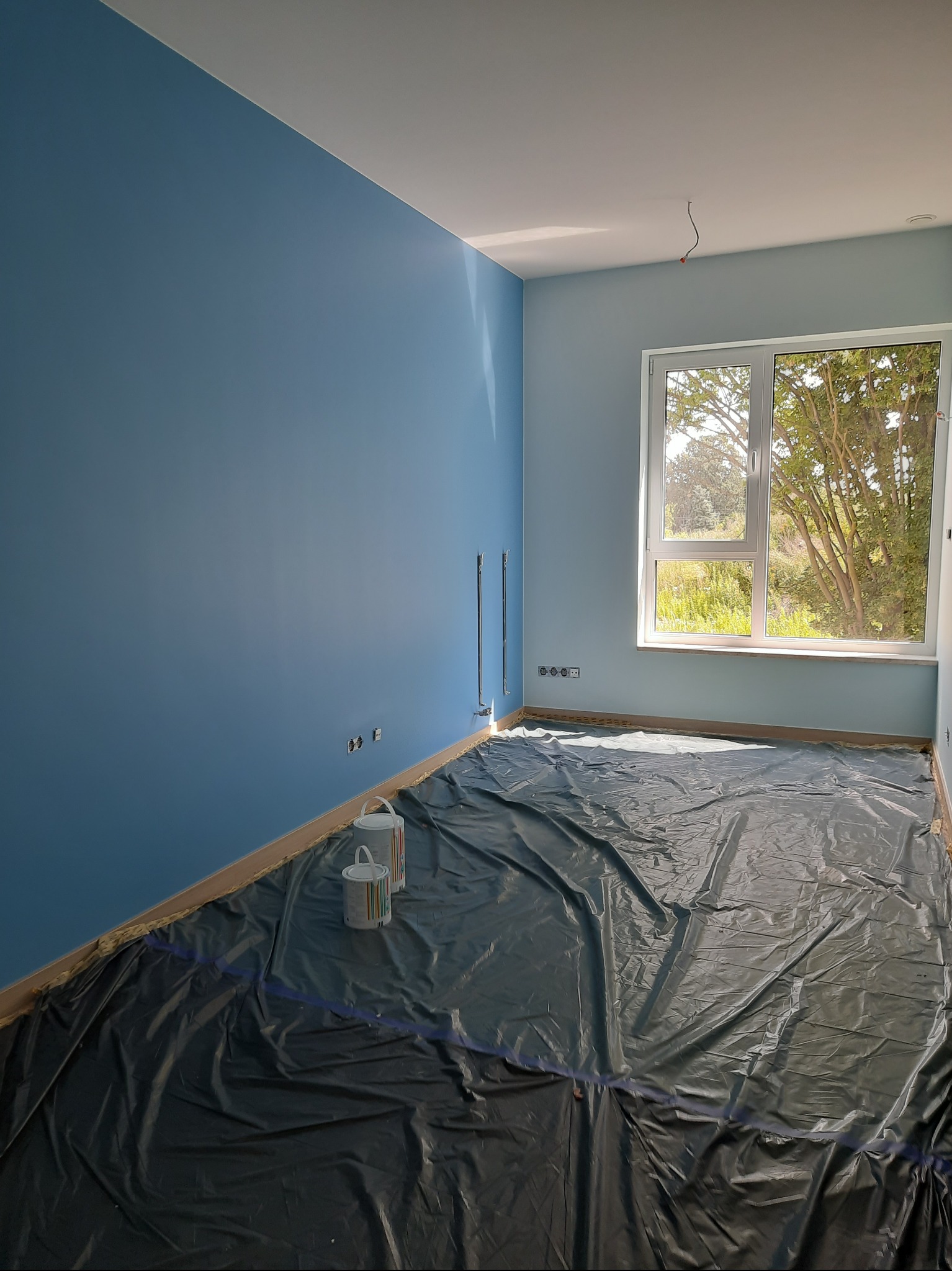 Pomieszczenie po malowaniu ścian na niebiesko, zabezpieczone folią i z puszkami farby. Widoczne gniazdka elektryczne i okno z widokiem na zieleń.