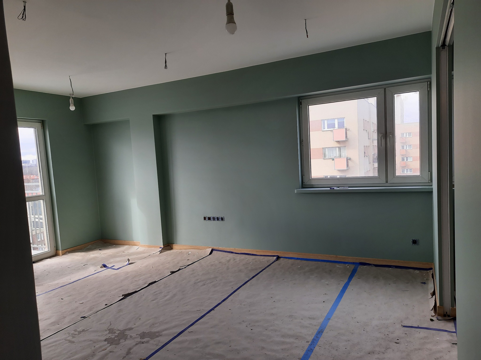 Pokój po malowaniu ścian na zielono, zabezpieczona podłoga folią i taśmą malarską. Widoczne gniazdka elektryczne i okno z widokiem na blok.